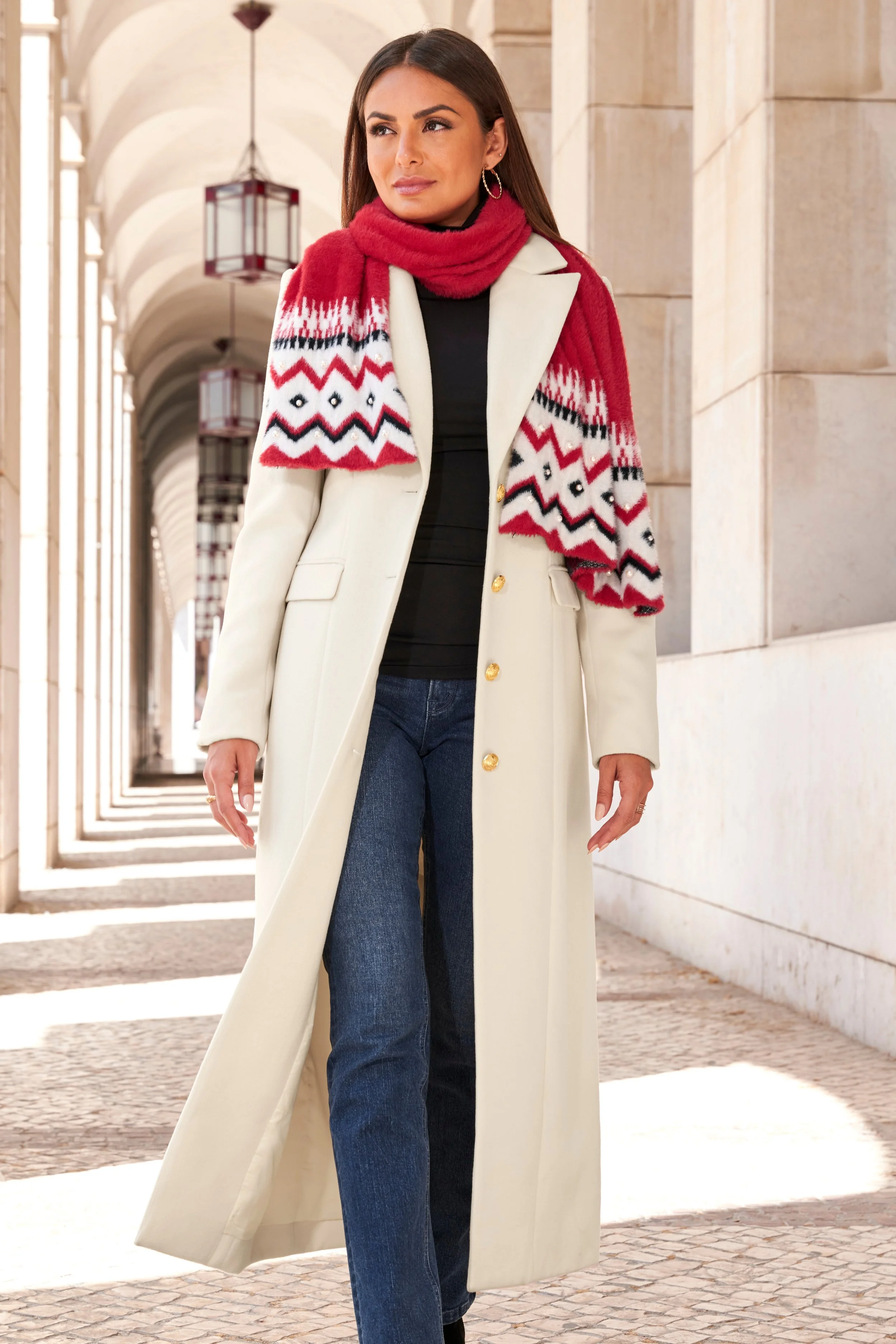 A woman with long dark hair walking outdoors in an arched walkway, wearing a white coat, red patterned scarf, black top, and blue jeans.