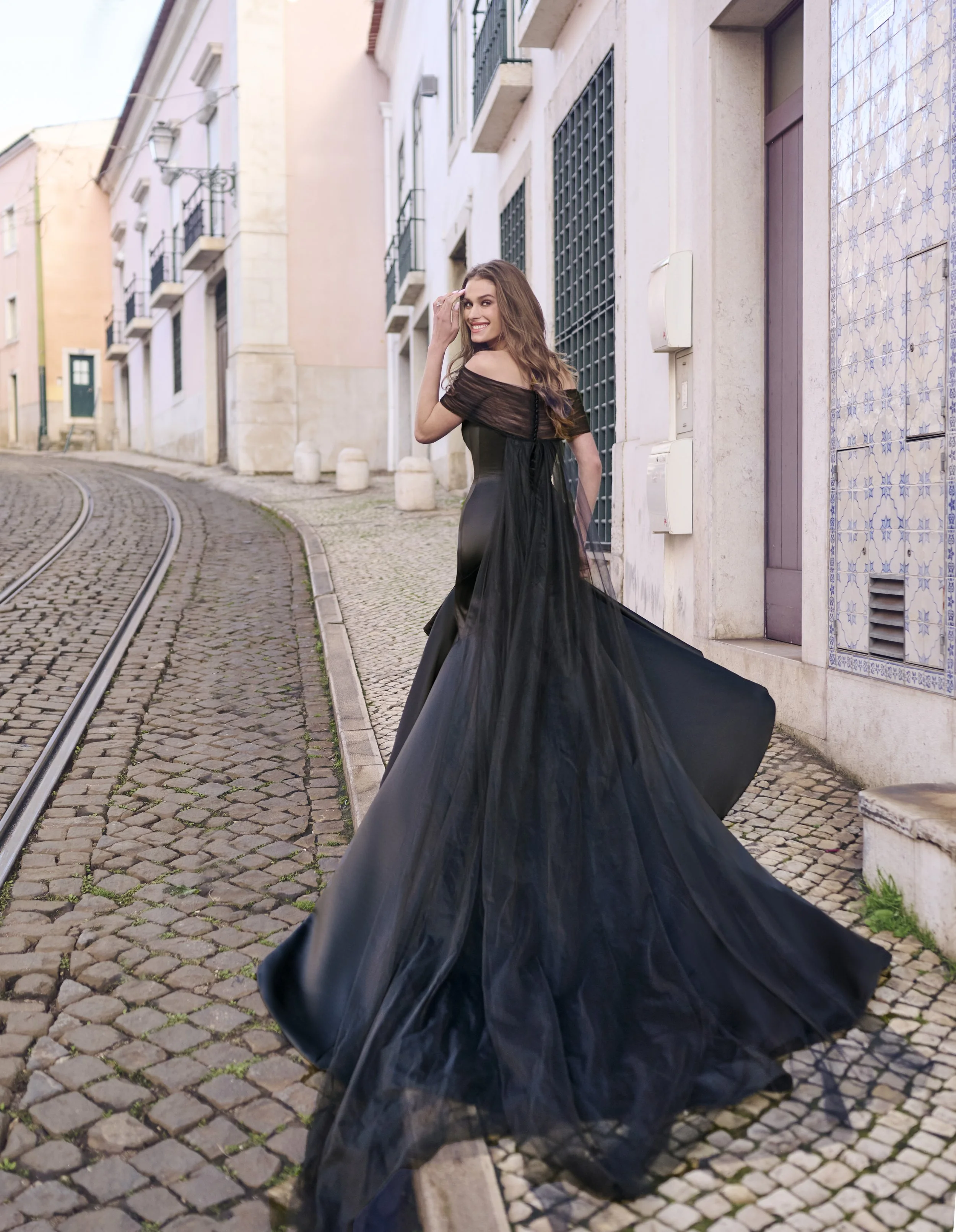 A woman wearing a black off-shoulder dress walking on cobblestone street with colorful buildings in the background.