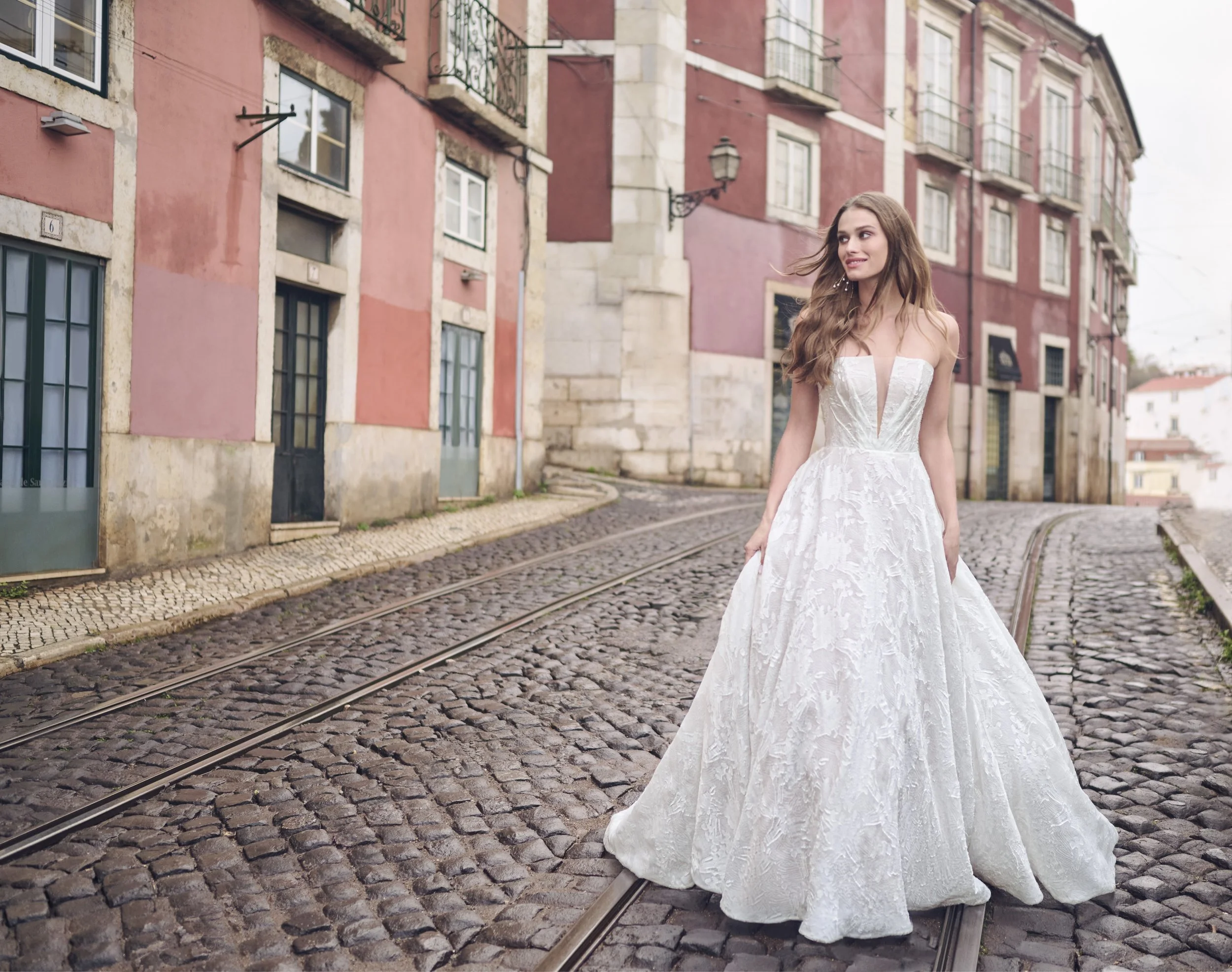 A woman in a white wedding dress with lace details, standing on cobblestone street near tram tracks, with pink painted buildings and windows with balconies in the background.