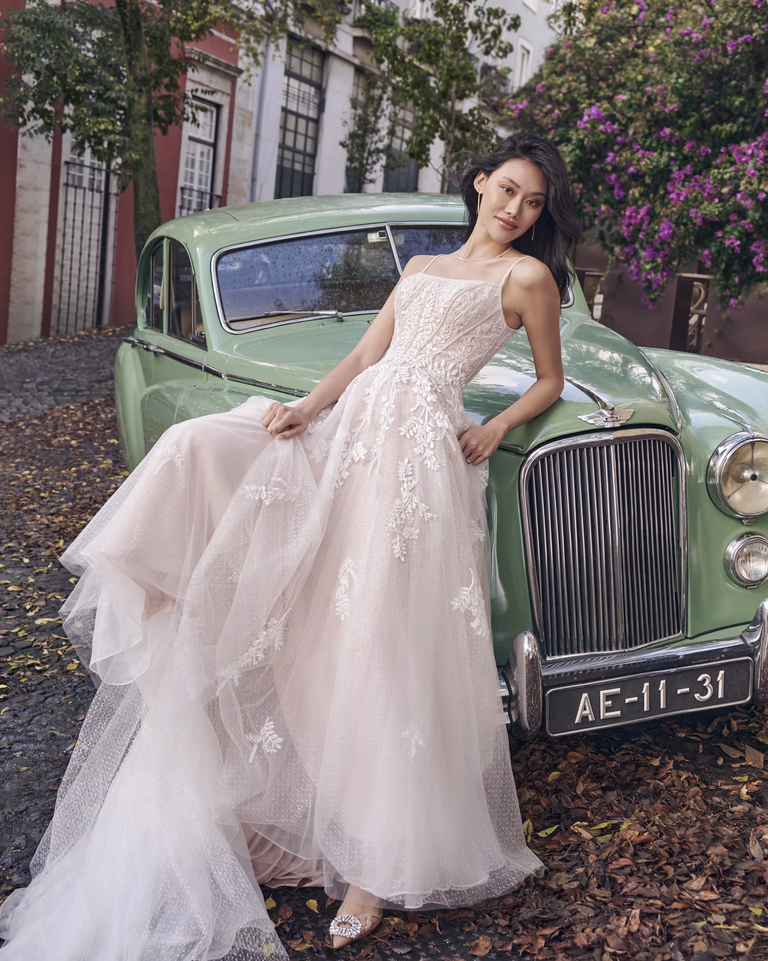 A woman in a light pink, elegant dress with lace details, standing in front of a vintage green car. She has long, wavy black hair and is wearing jewelry. The background features trees, purple flowering bushes, and buildings with closed windows.