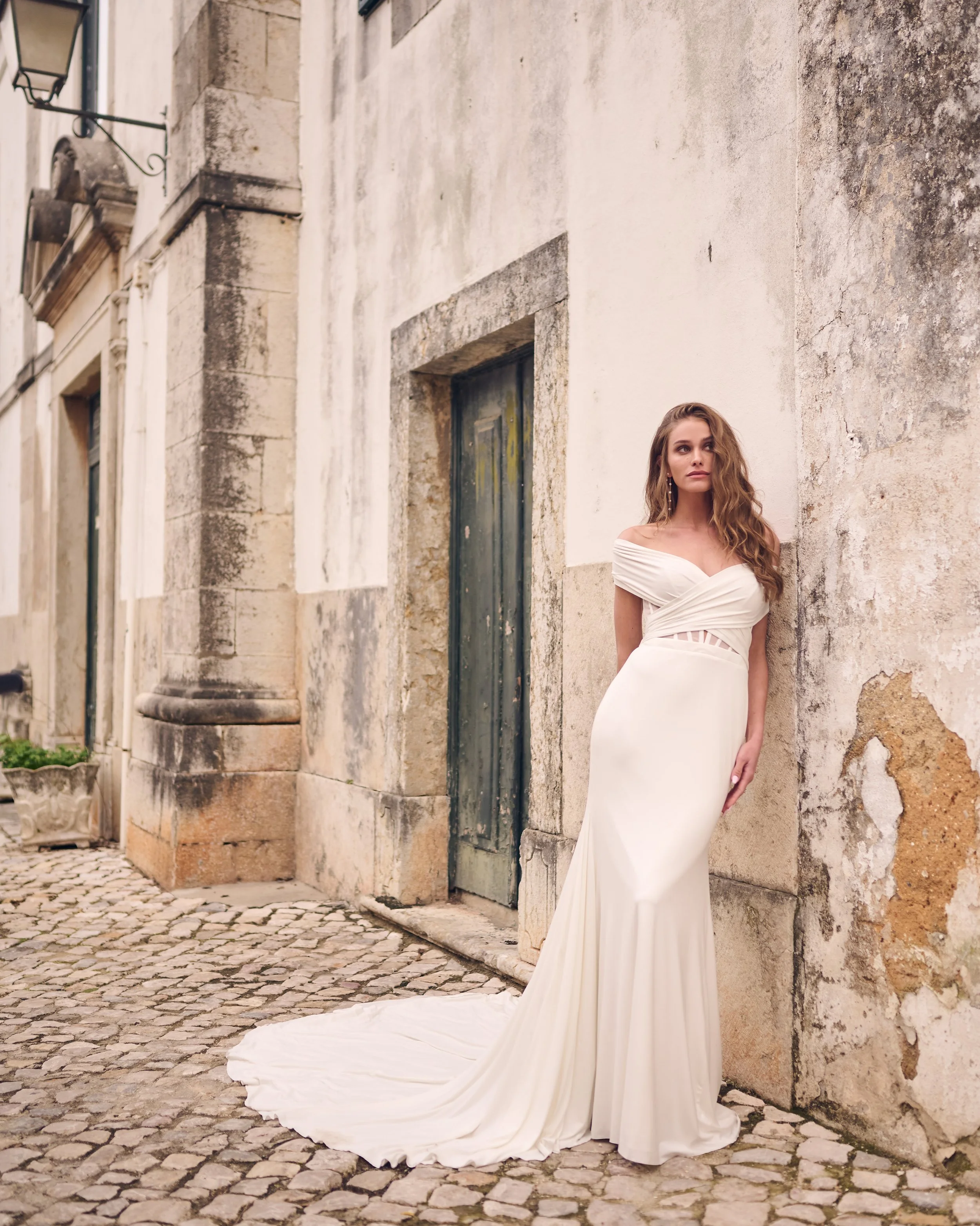 A woman in a white off-shoulder gown with a long train standing against an aged stone wall in a cobblestone street, outdoors.