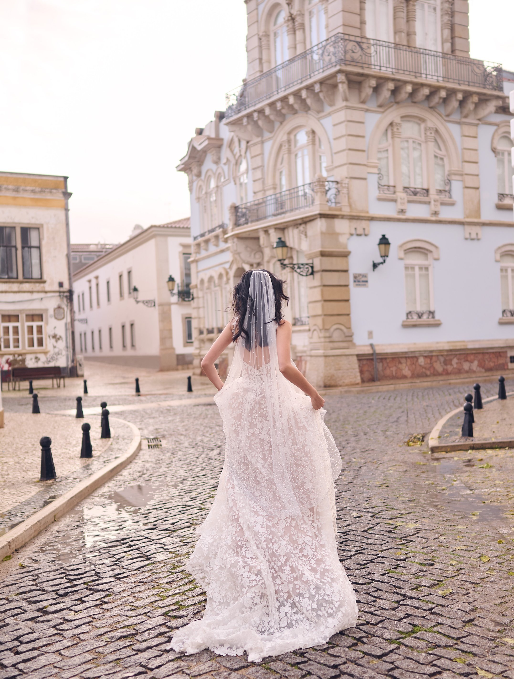 A woman wearing a wedding dress and a veil stands on a cobblestone street in front of ornate buildings with balconies and street lamps.