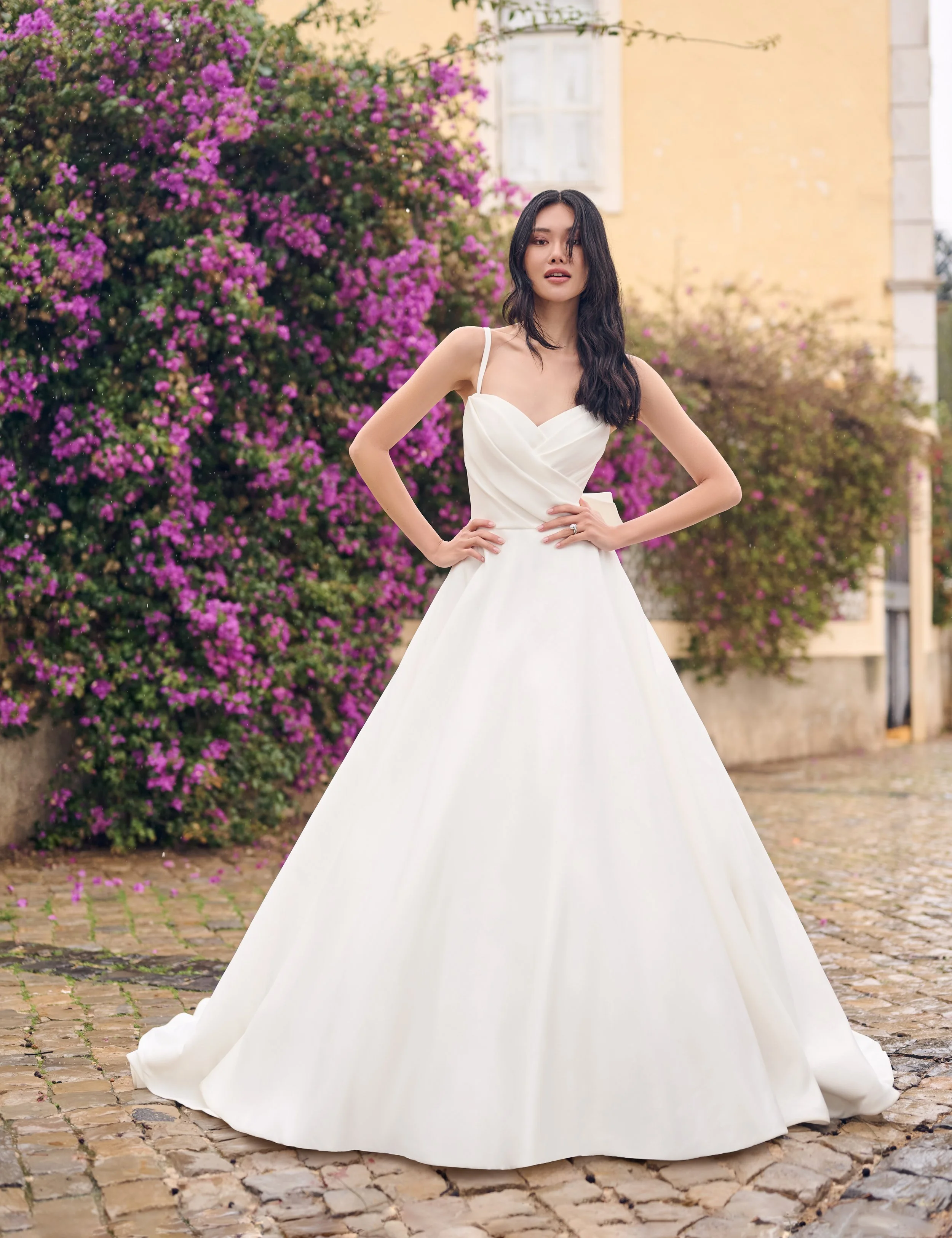A woman in a white wedding dress standing on a cobblestone street with pink flowering bushes behind her.