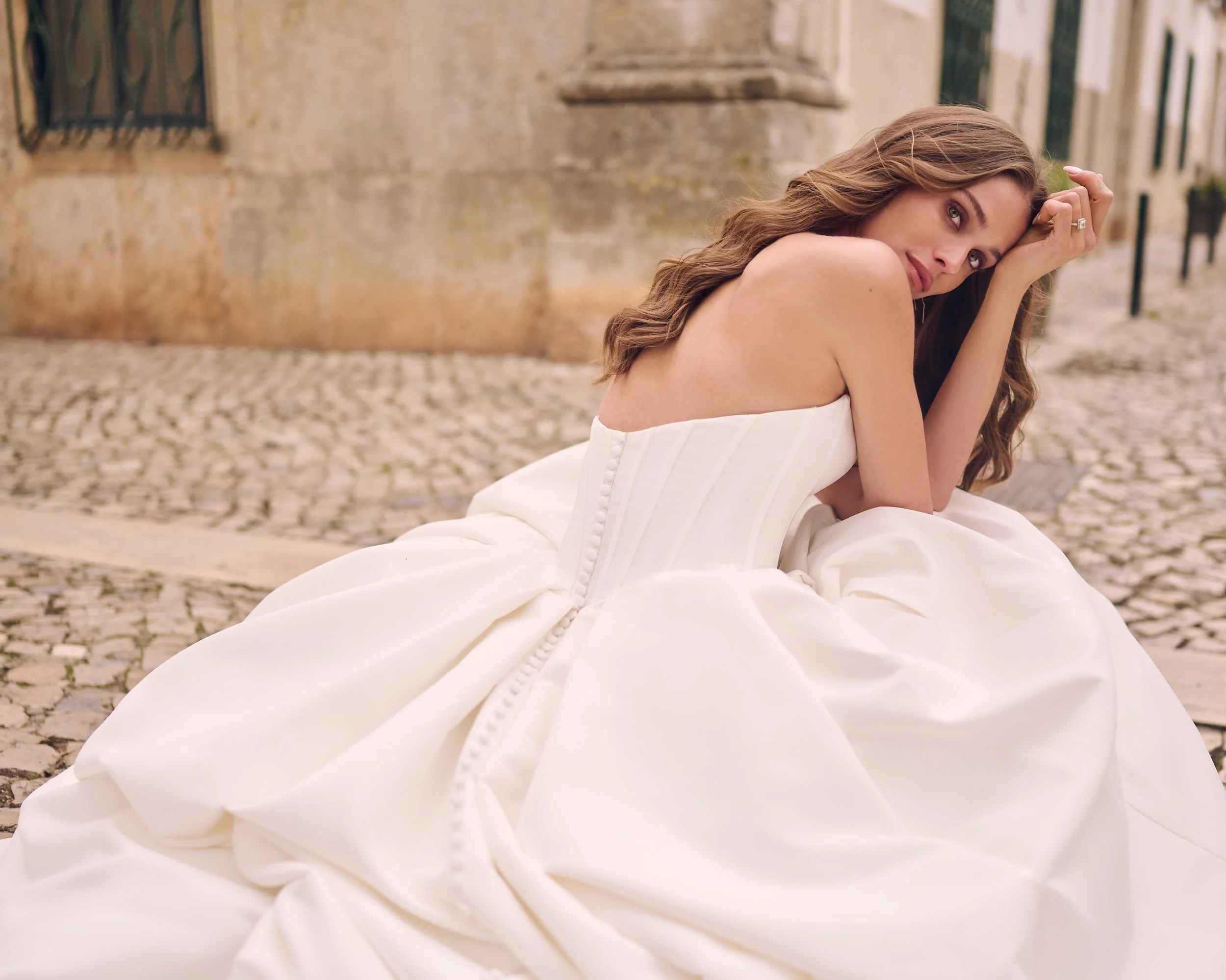 A woman with wavy brown hair and wearing a white strapless dress sitting on cobblestone street, resting her head on her hand, with older stone buildings in the background.