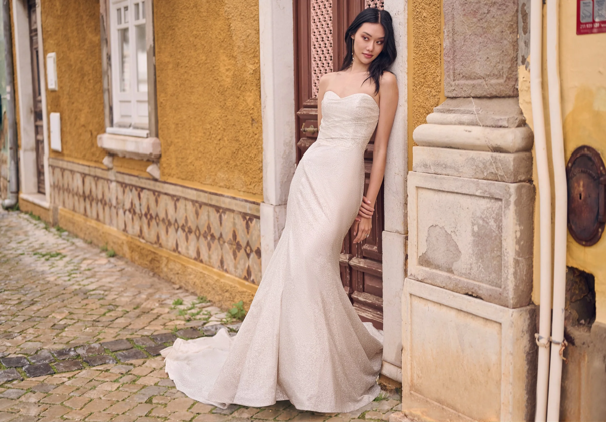 A woman in a strapless, elegant beige gown leaning against an ornate stone wall, with a colorful building and cobblestone street in the background.