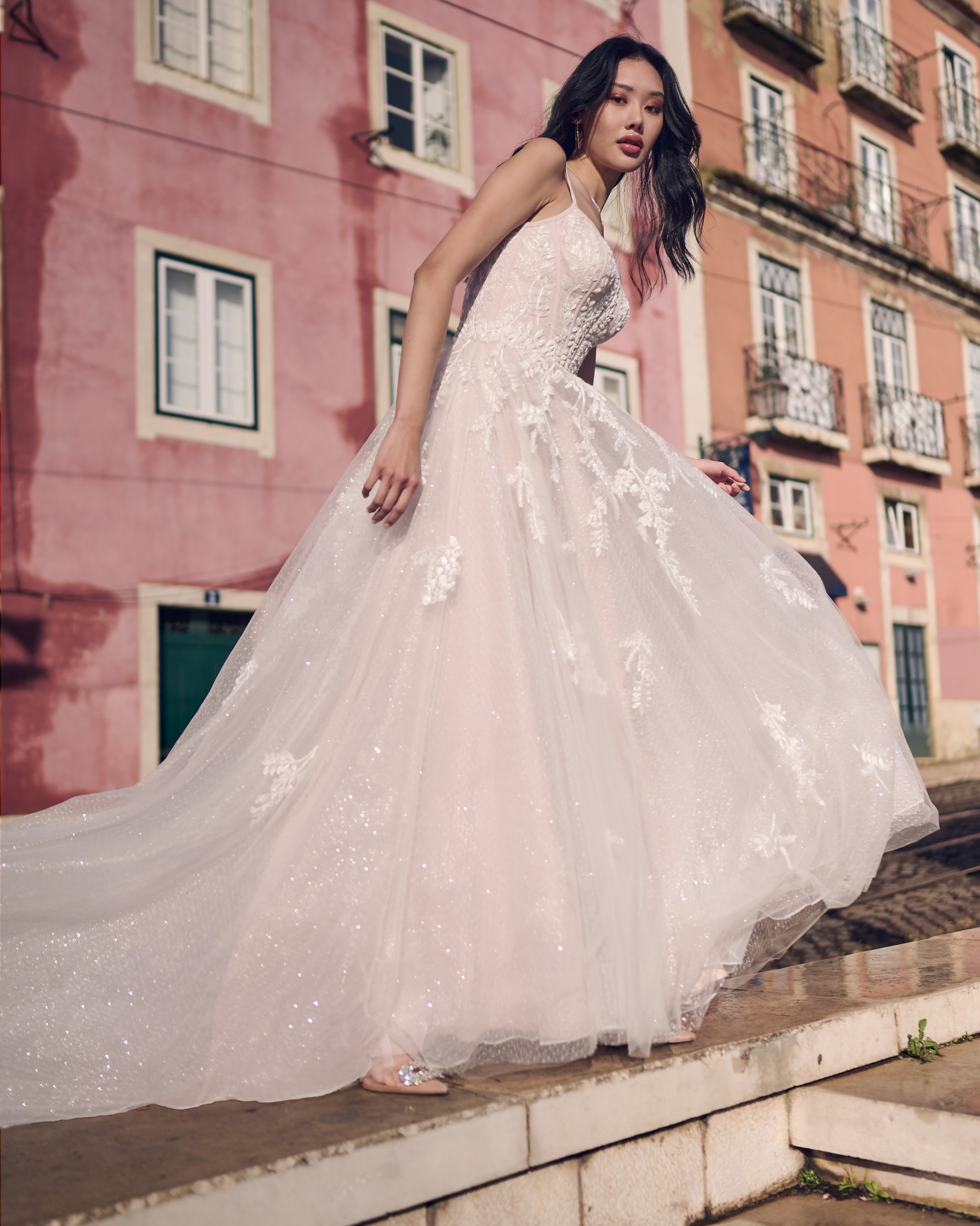 A woman in a white wedding dress with lace and floral embroidery, standing on a stone staircase in front of a pink building with balconies and multiple windows.