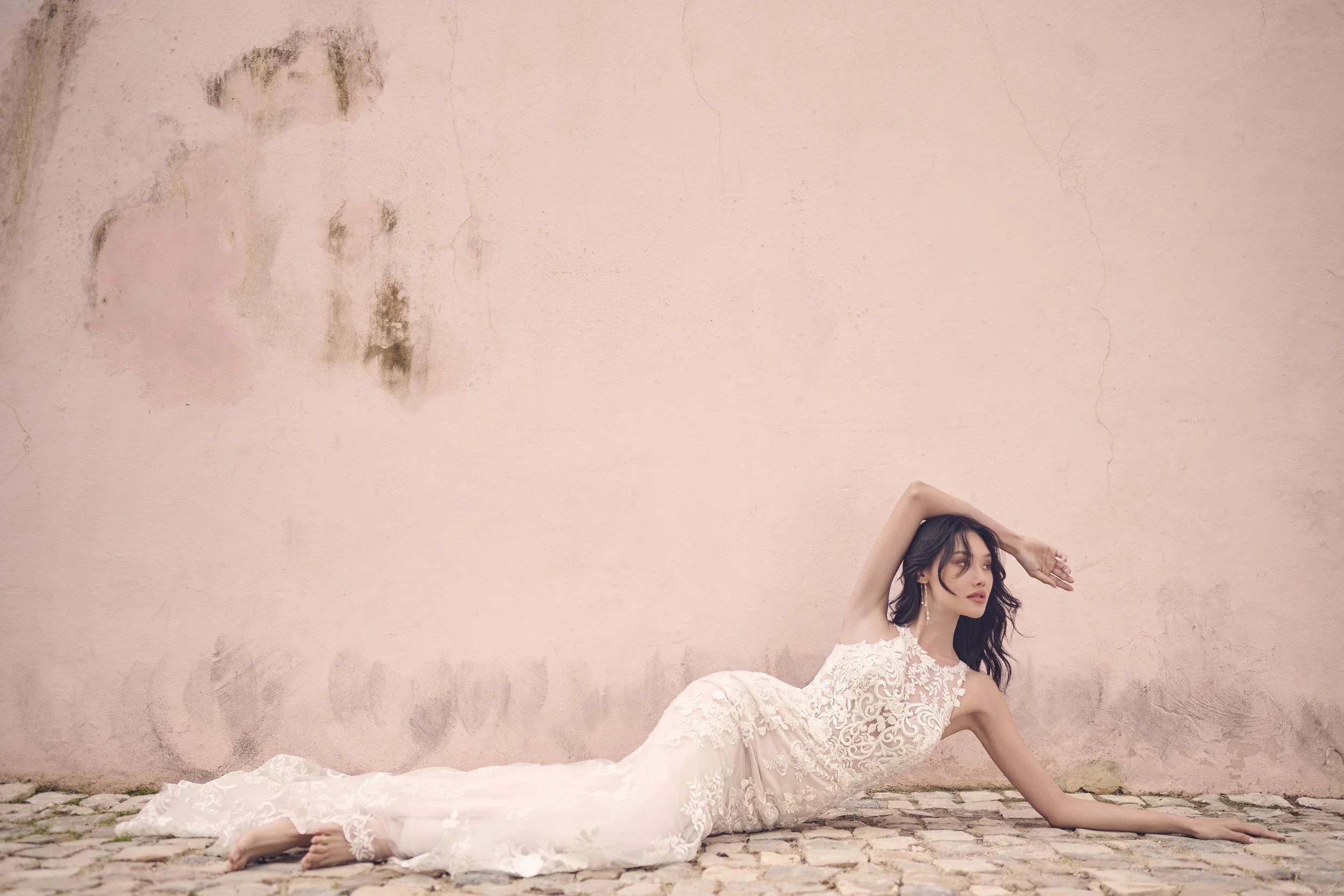 A woman in a white lace dress lying on the cobblestone ground, her head resting on the ground and her hand on her head, with a pink wall behind her.