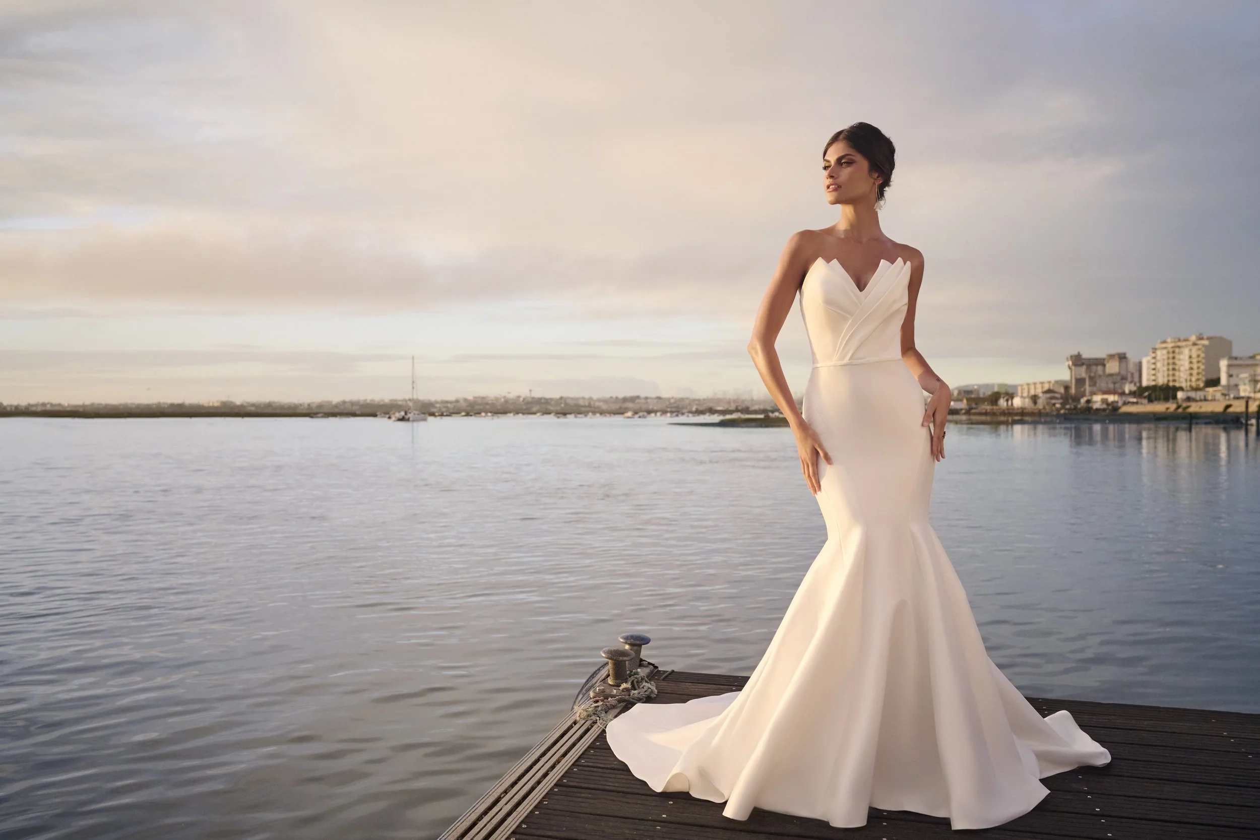 A woman in a white strapless wedding gown stands on a dock by the water, with a city skyline and buildings in the background, during sunset.