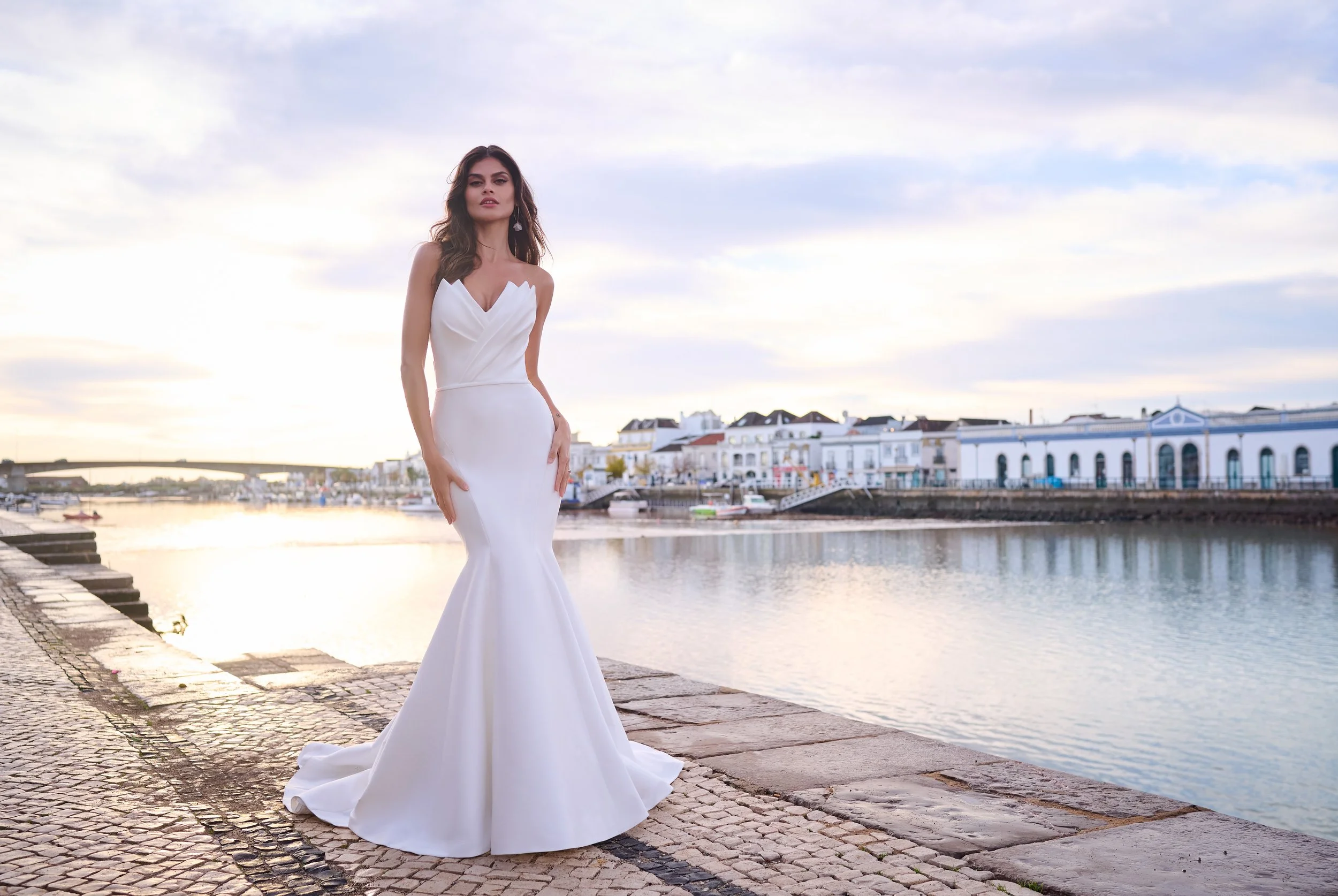 A woman in a white wedding gown standing on a stone pathway by the water, with a background of pastel-colored buildings and a cloudy sky.