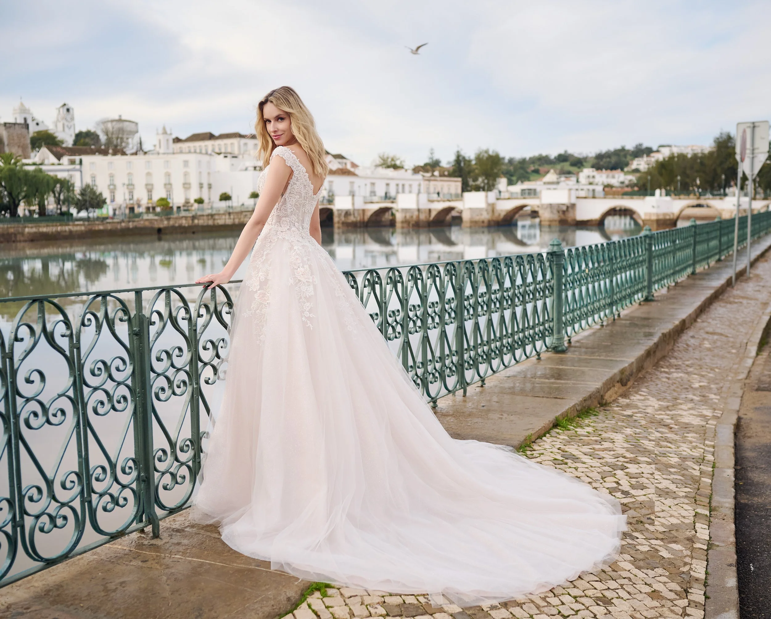 A woman in a white lace wedding dress with a long train standing by a decorative metal railing by a river, with white buildings and a bridge in the background.