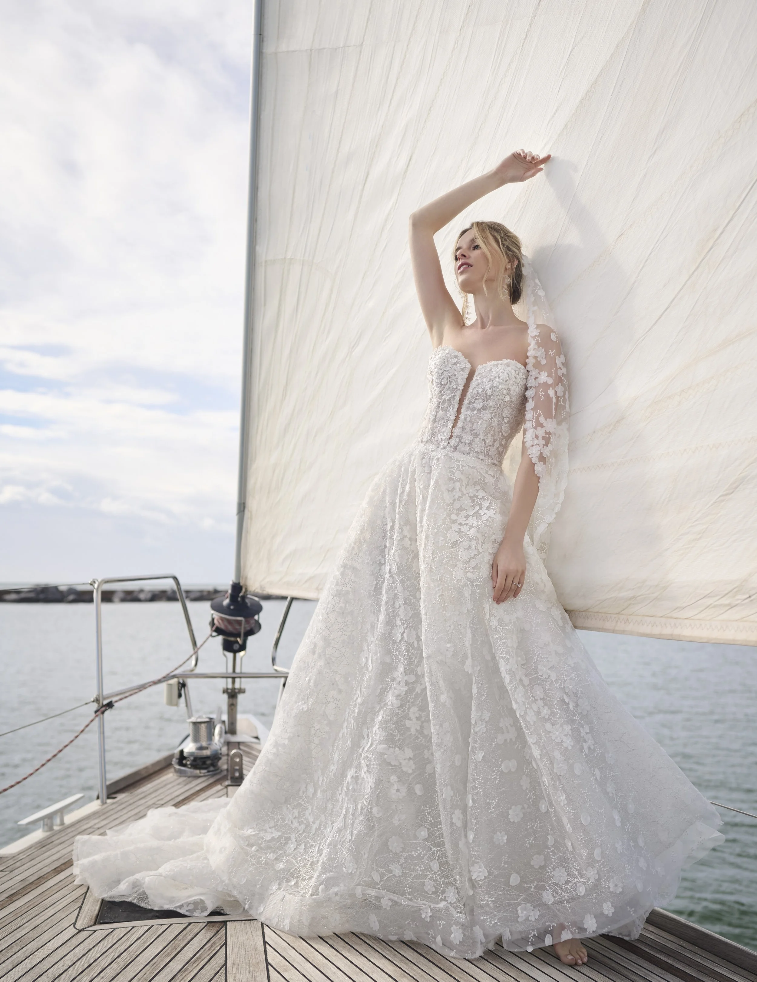 A woman in a white wedding dress with lace flower details standing on a boat dock, holding onto a sailboat's side with water and sky in the background.