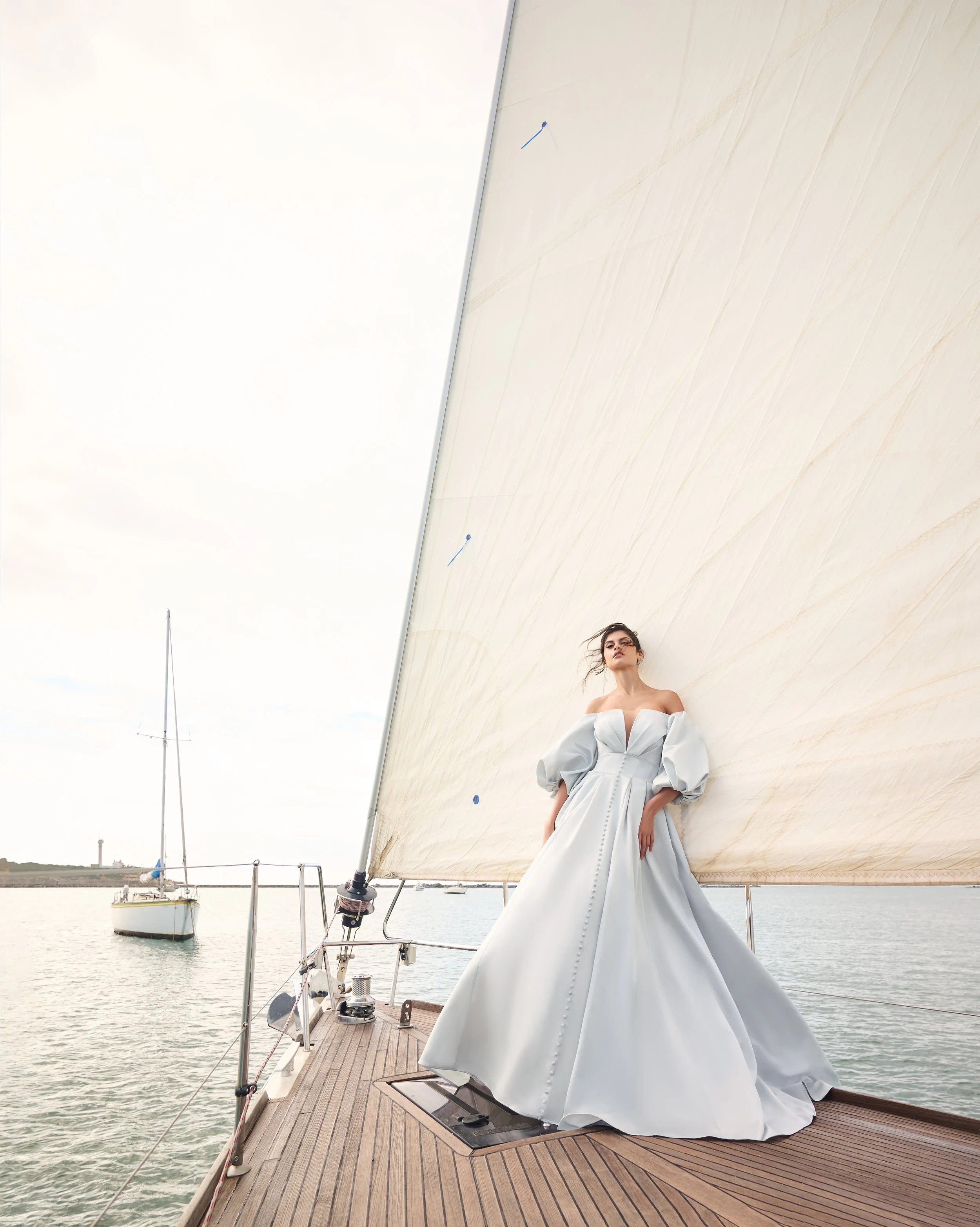 A woman in a white dress standing on the deck of a yacht with a beige sail in the background and water with boats in the distance.