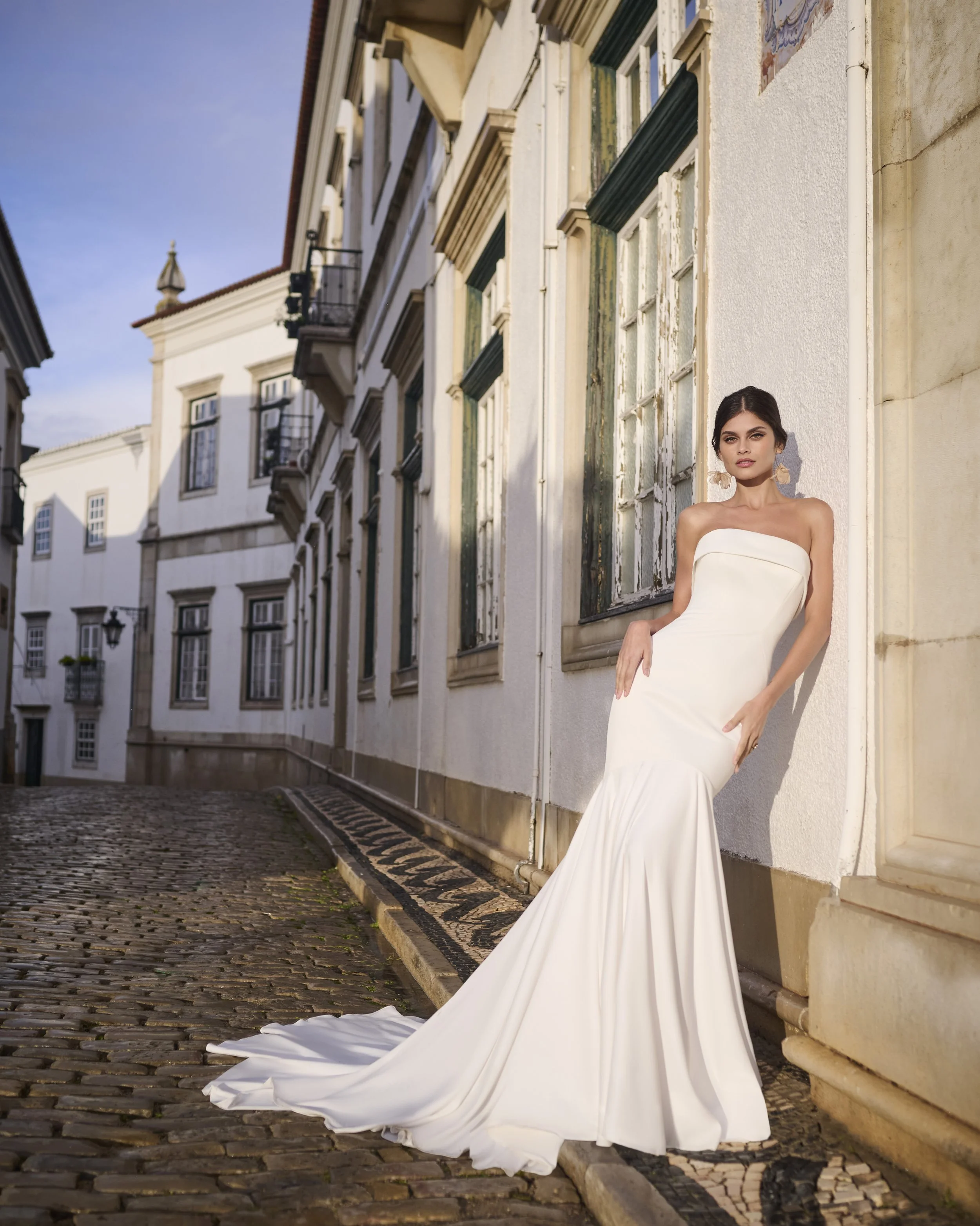 A woman in an elegant white strapless gown with a long train posing against a white building with tall windows and a cobblestone street, in a European city setting during daytime.