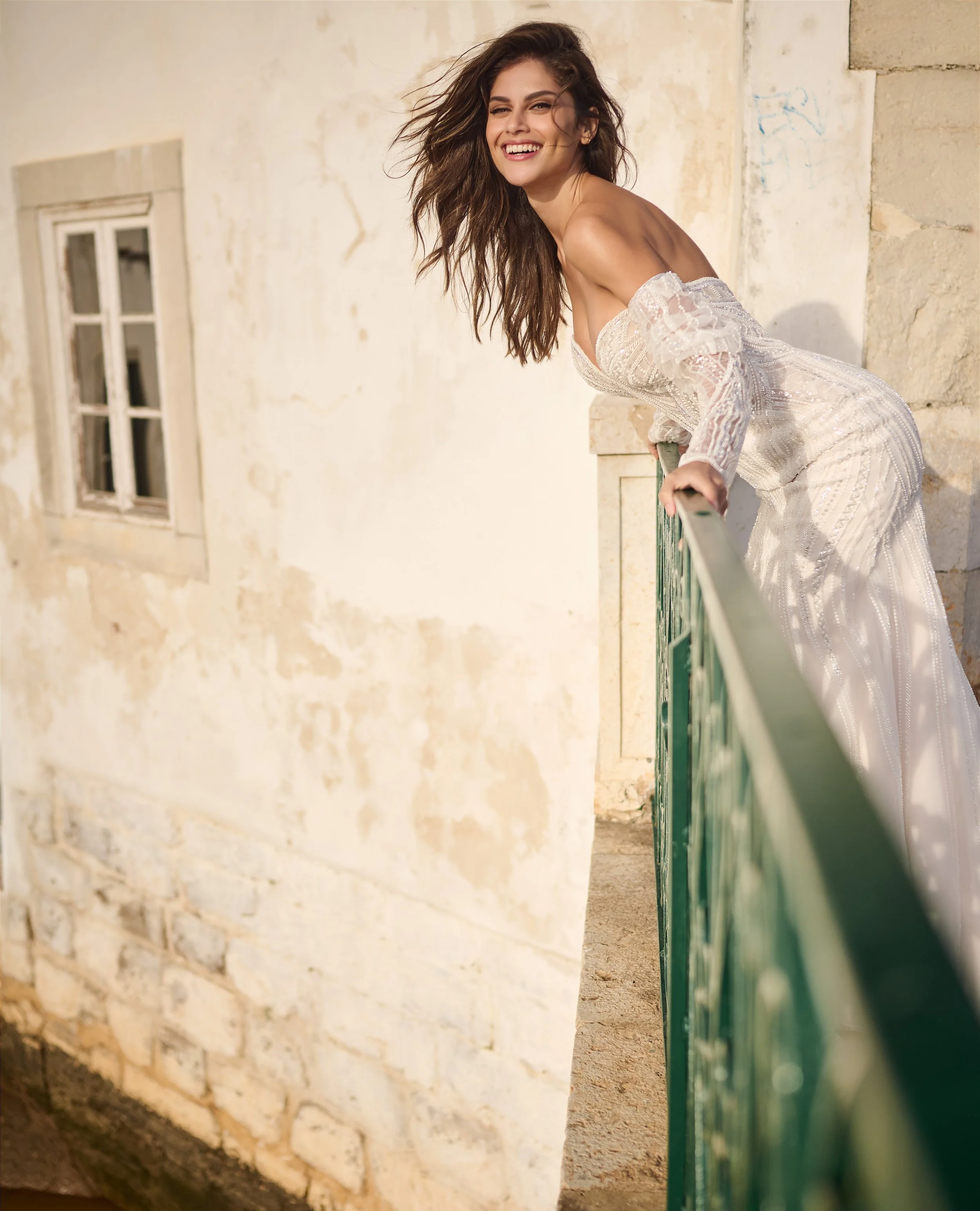 A young woman with long, wavy brown hair smiling and looking at the camera, leaning on a green railing outside a weathered, beige stone building with a window.