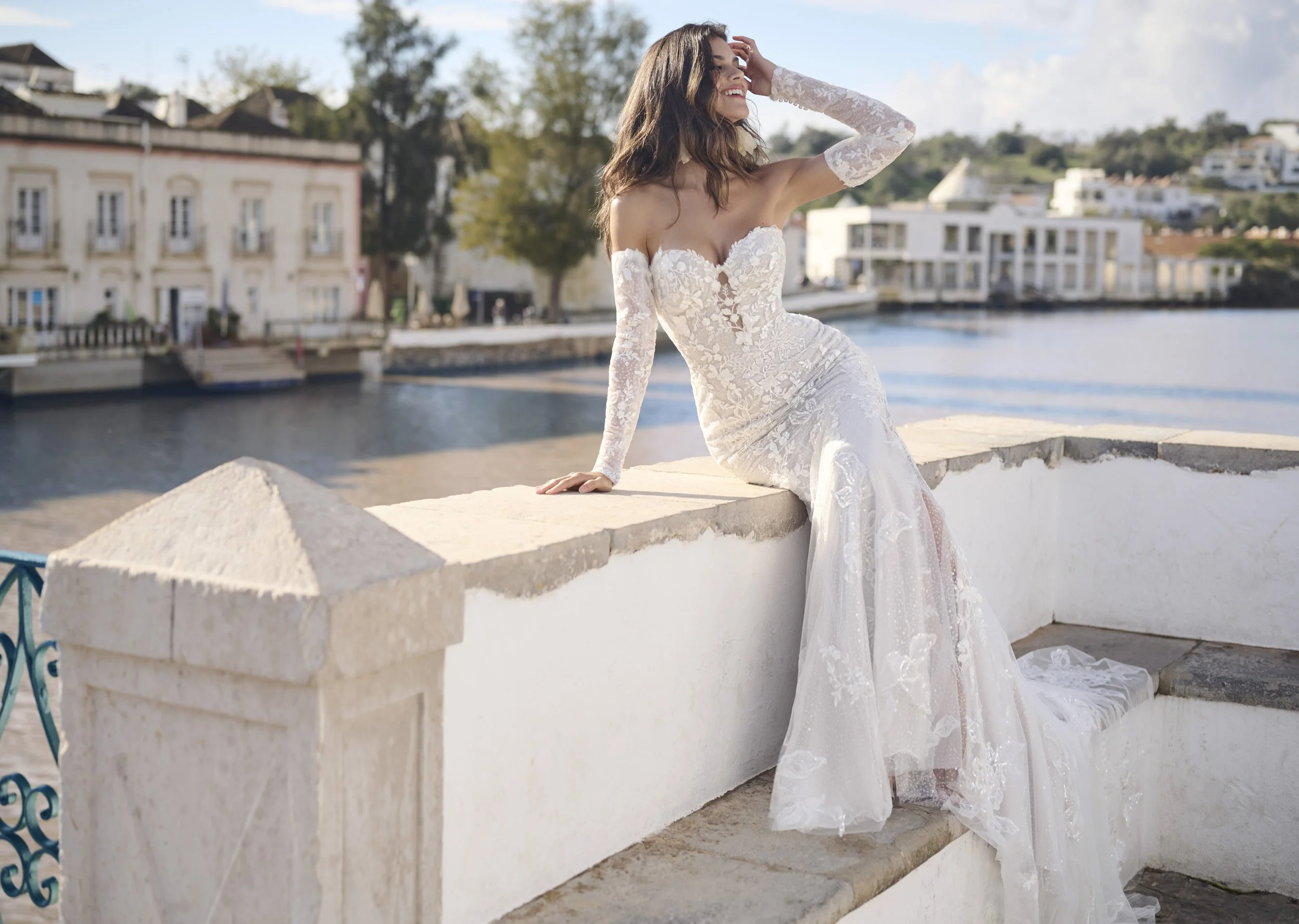 A woman in a white lace wedding gown with long sleeves, smiling and posing outdoors by a waterfront during daytime.