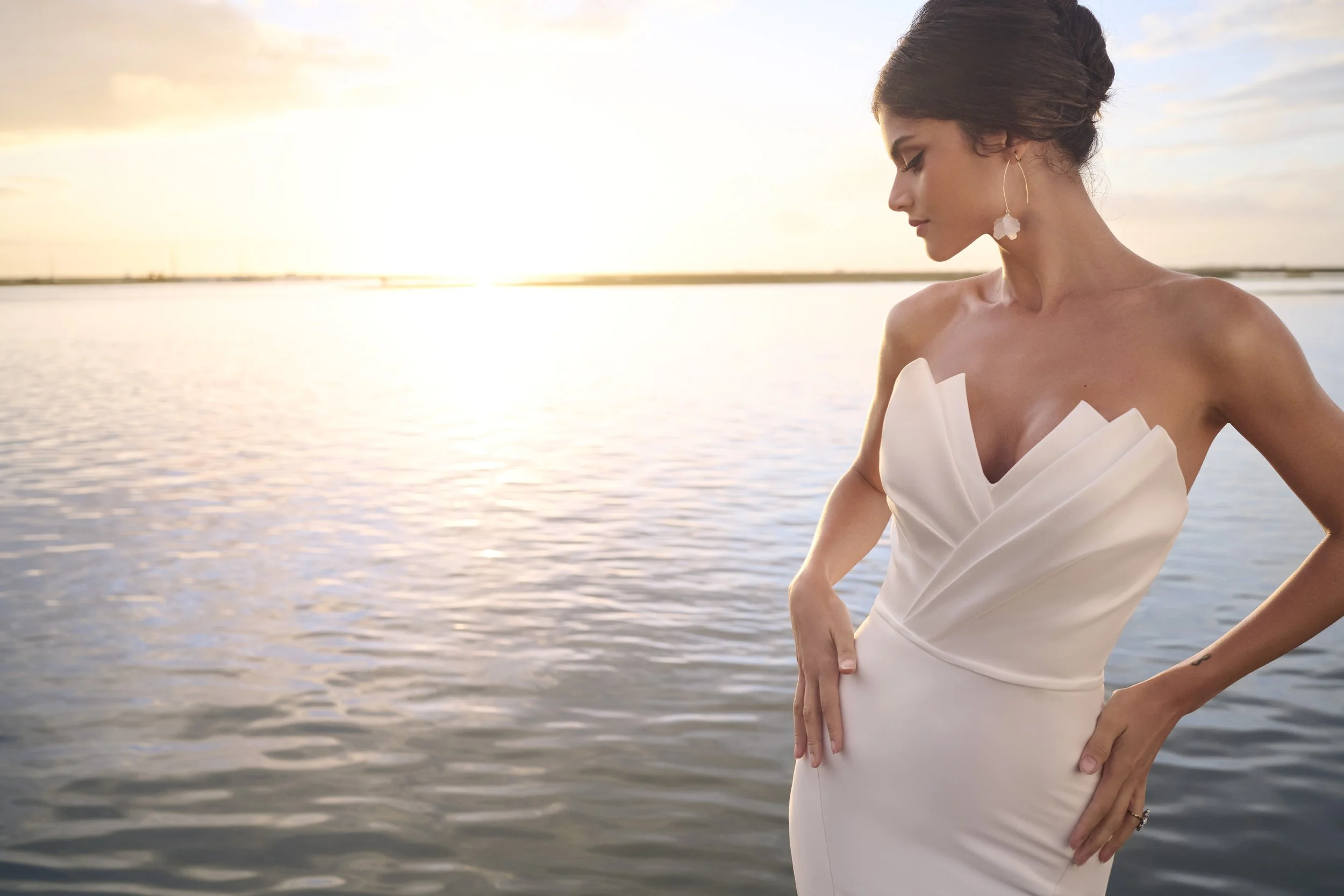 A woman in a white dress standing by a body of water during sunset, wearing a large earring and looking down with one hand placed on her stomach.