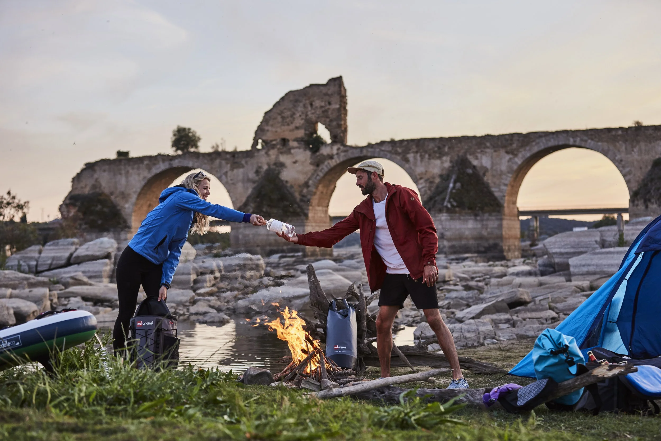 Two people, a woman in a blue jacket and a man in a red jacket, are camping near a river with a historic stone bridge in the background. They are toasting cups over a small campfire.