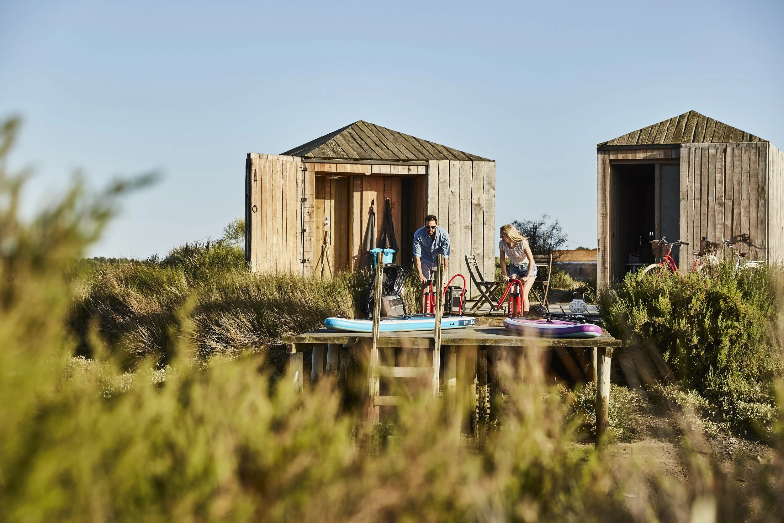 A man and woman are preparing a fire outside two wooden beach huts near the water, with paddleboards on the dock and bicycles nearby, on a clear sunny day.