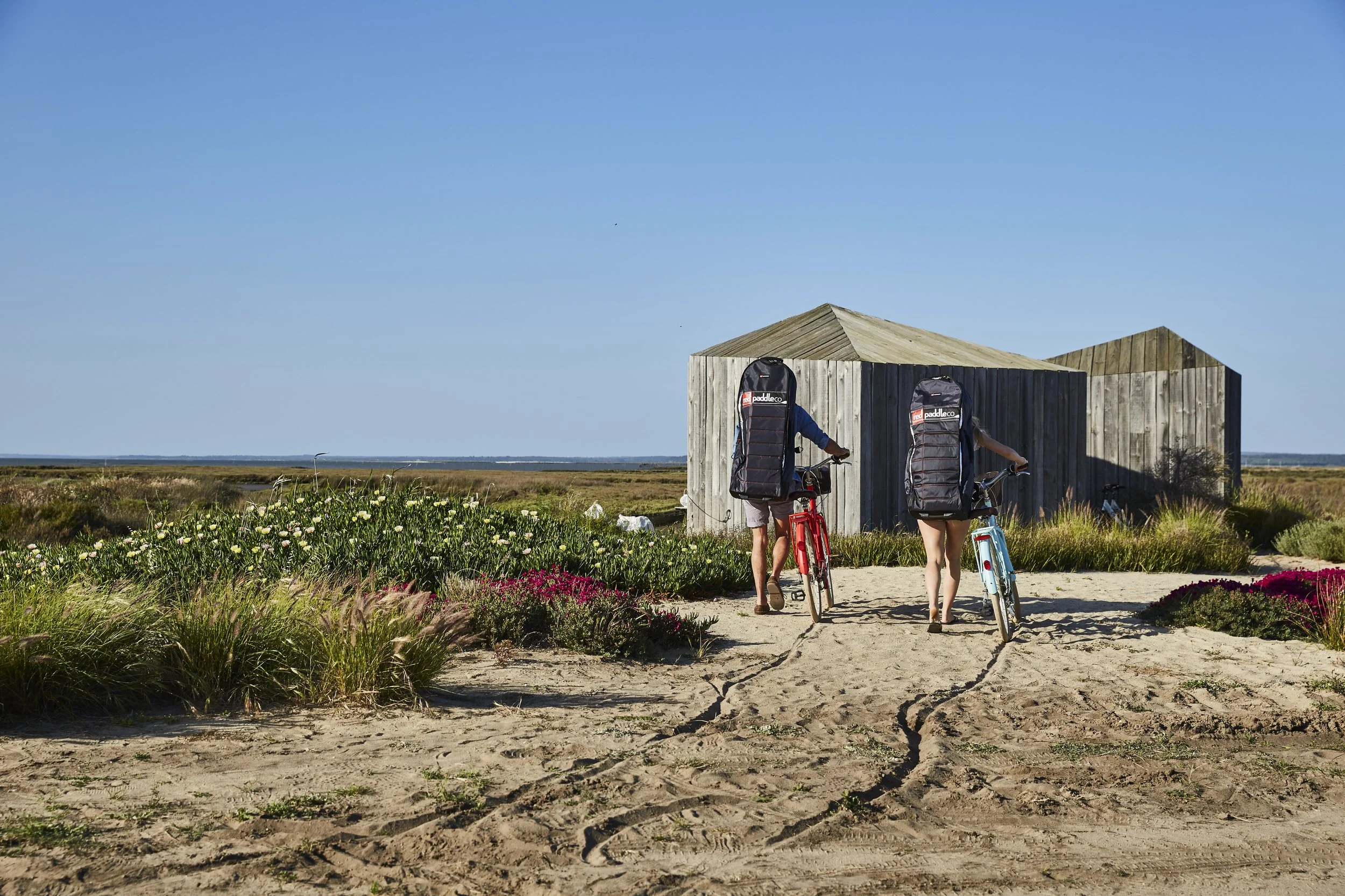 Two people with large backpacks on bicycles walking toward a wooden beach hut on a dirt path, with flowers and vegetation in the foreground and a blue sky in the background.