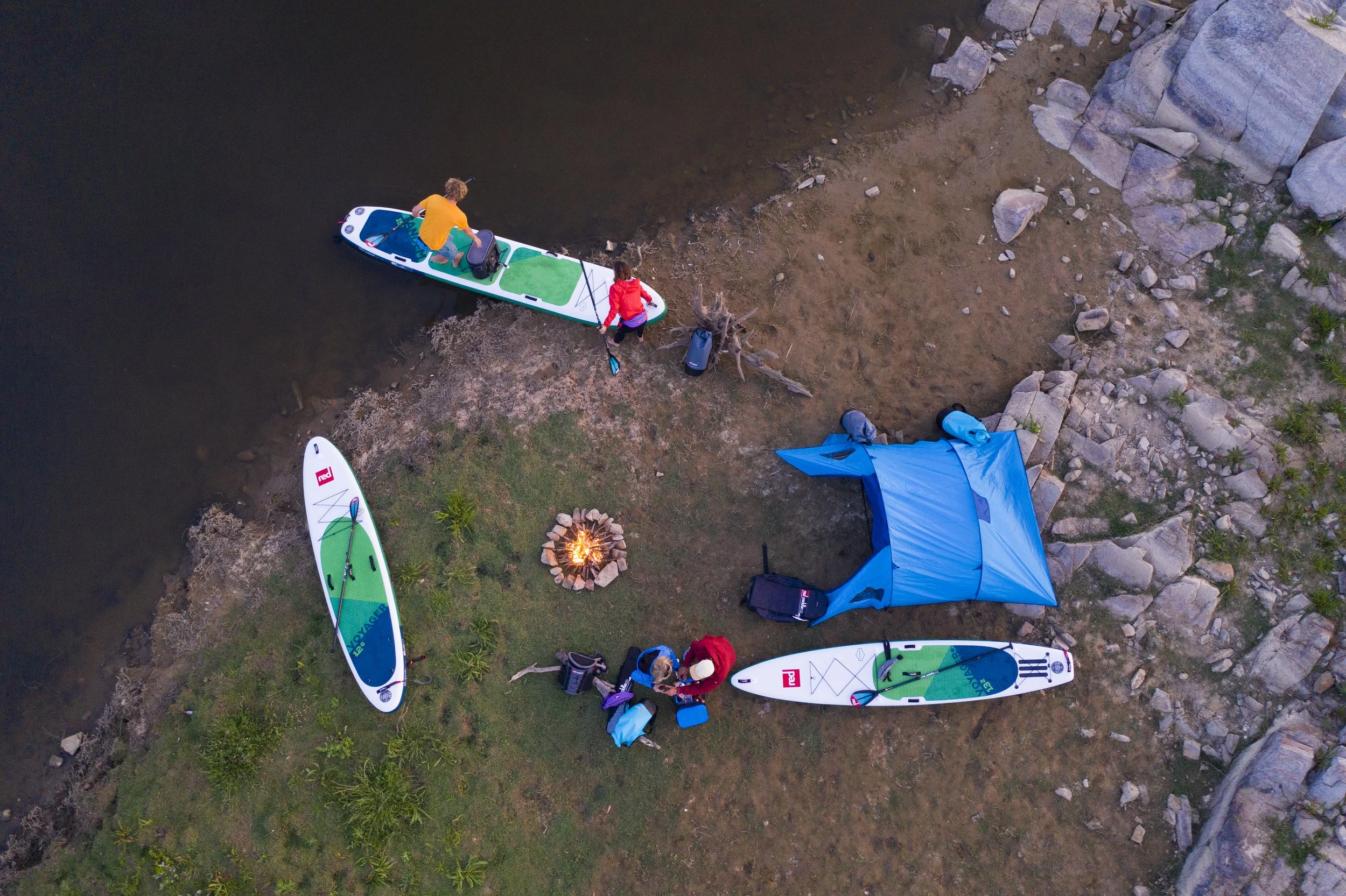 An aerial view of two people preparing paddleboards on a grassy area next to a rocky shoreline, with a campfire burning surrounded by stones and a blue tent nearby.