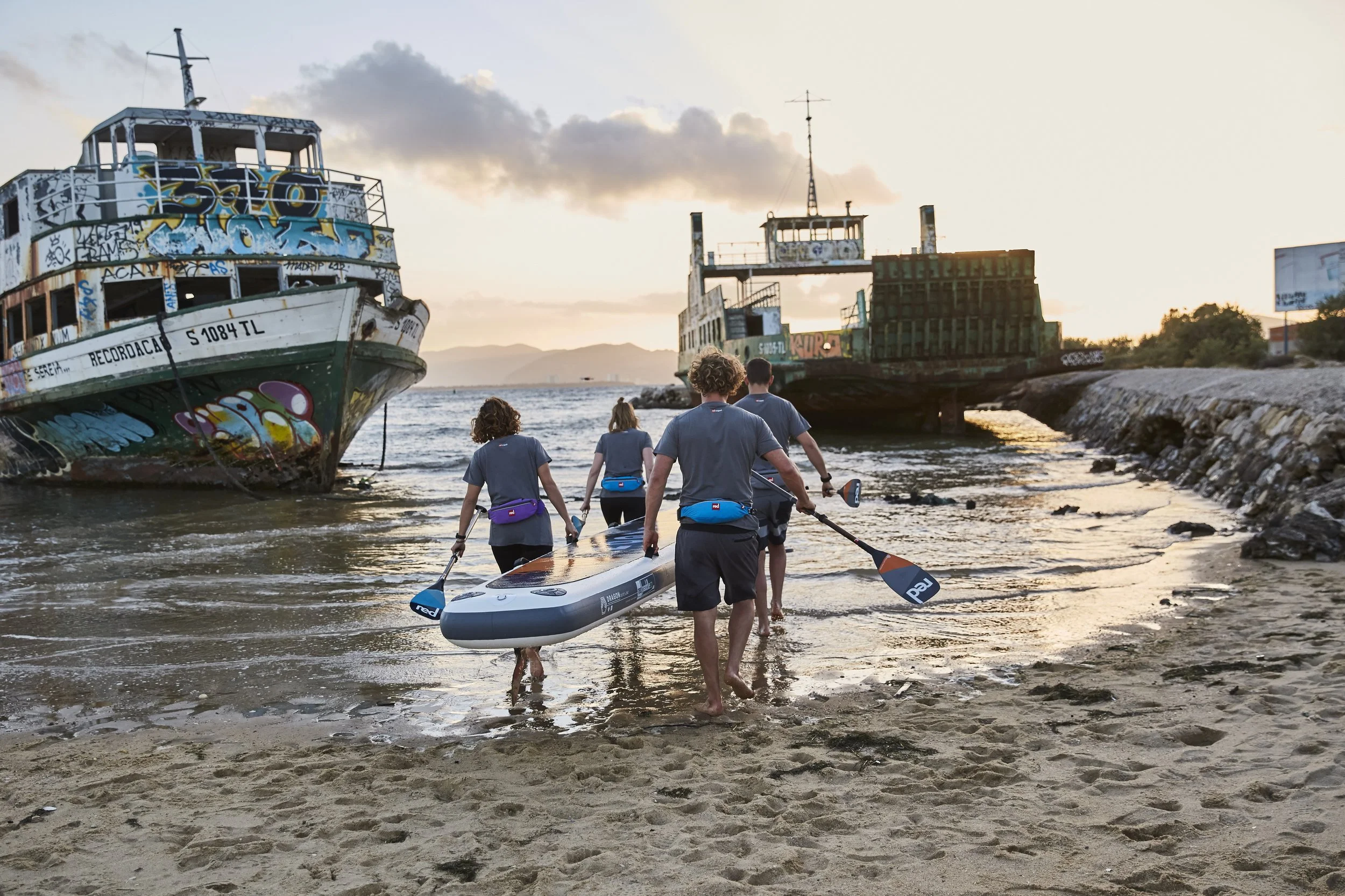 A group of five people wearing life jackets carrying a paddleboard into the water, with two abandoned graffiti-covered boats on the shore and a sunset sky in the background.
