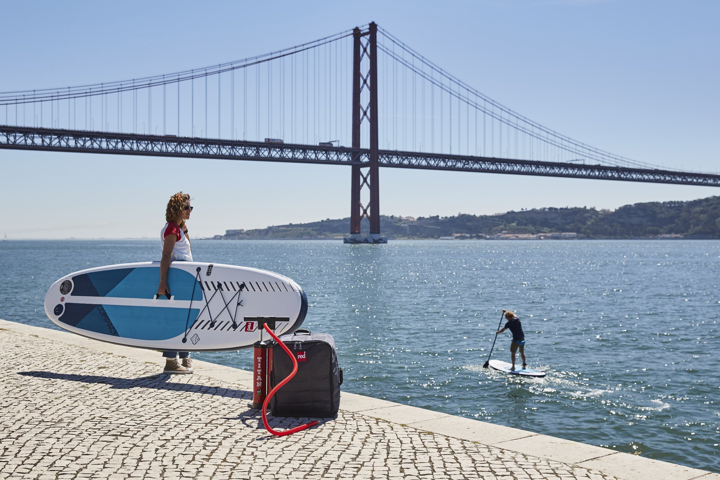 Woman with curly hair wearing sunglasses holding a stand-up paddleboard on a paved waterfront promenade with a bridge and water in the background. A person paddleboarding on the water with a dog nearby.