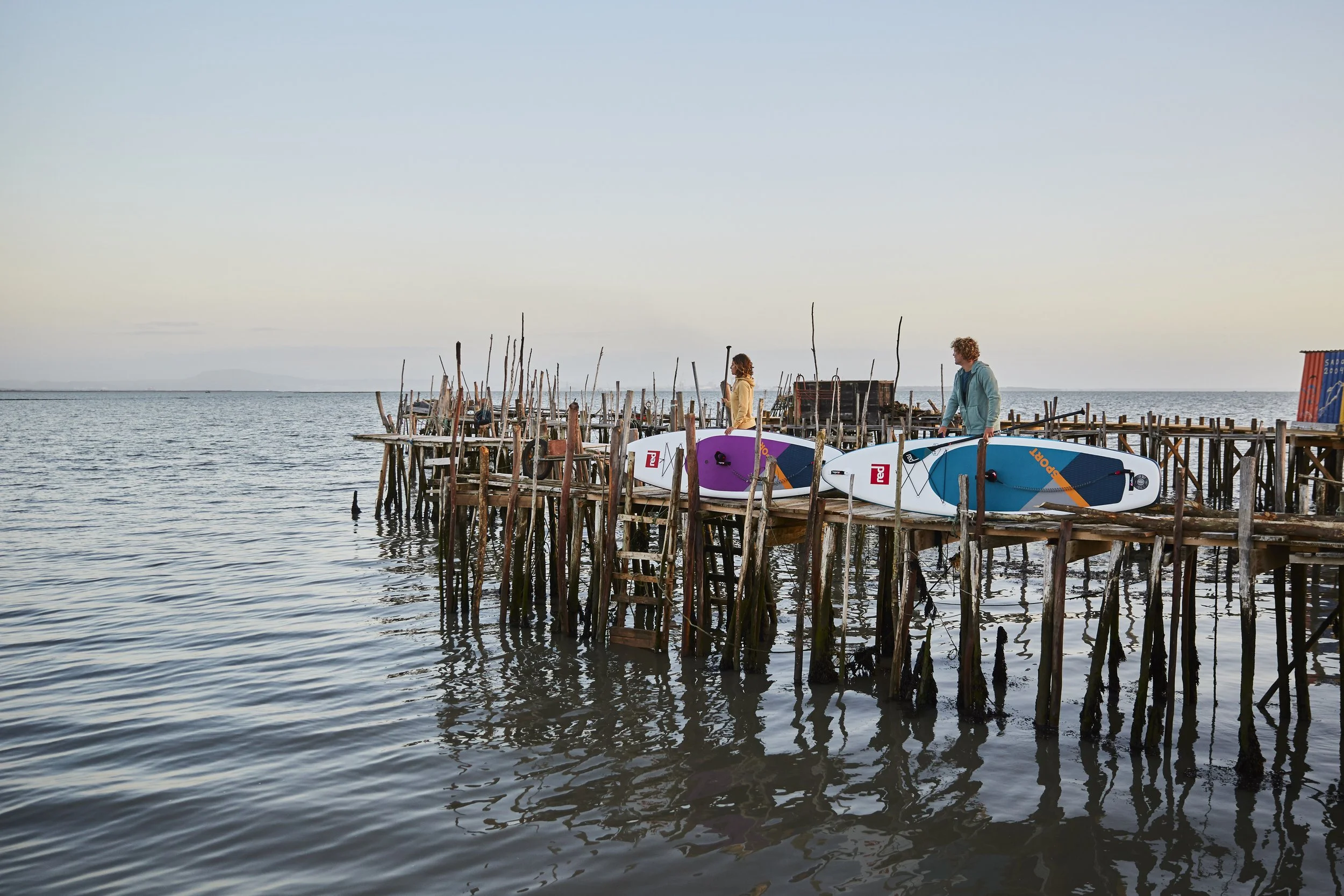 A wooden dock extending into a body of water with two people standing on it, each holding a paddleboard, at sunset or dawn with calm waters and a clear sky.