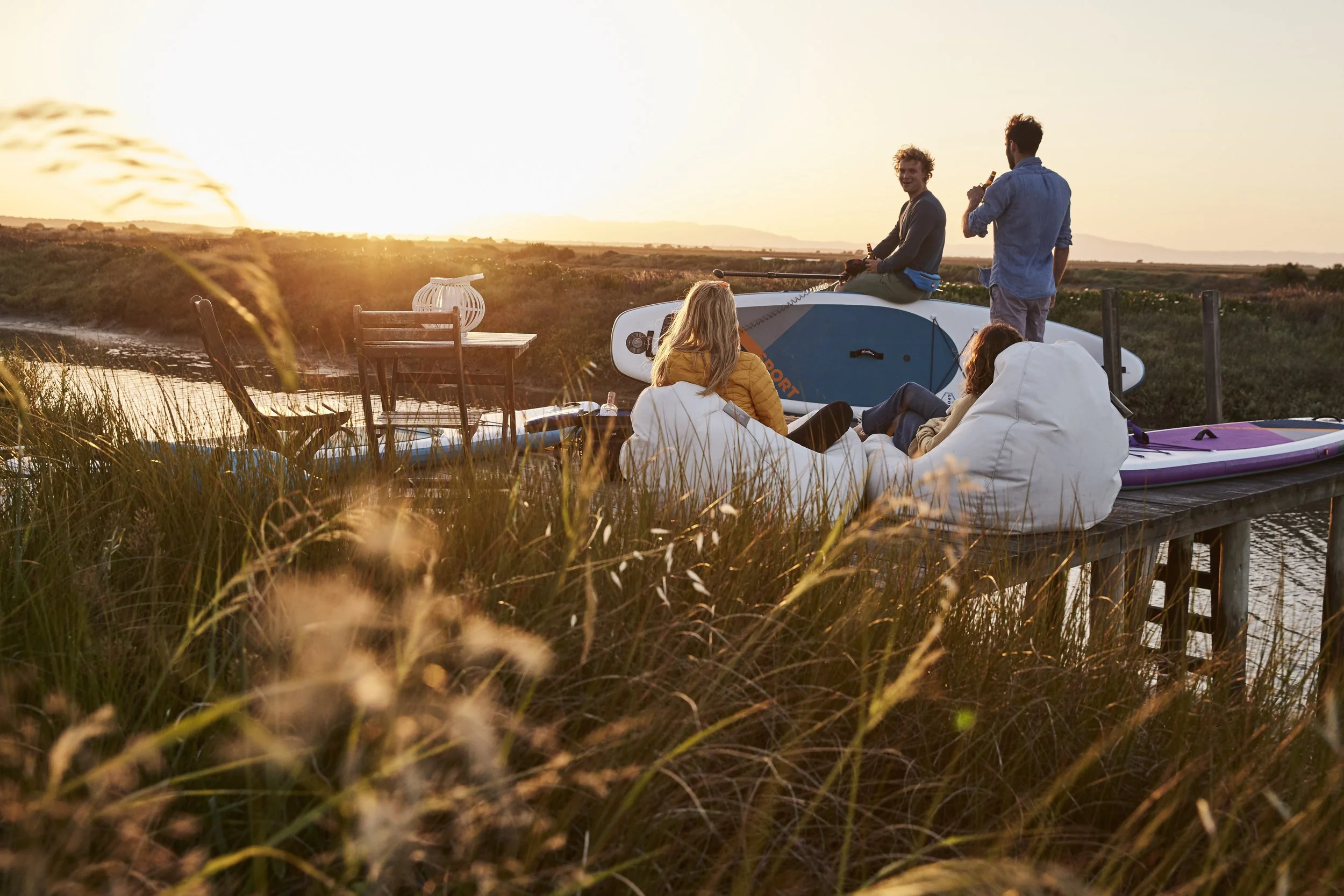 Group of friends relaxing by a waterway during sunset, with one person sitting on a paddleboard and others sitting on bean bags or chairs, enjoying drinks and conversation.