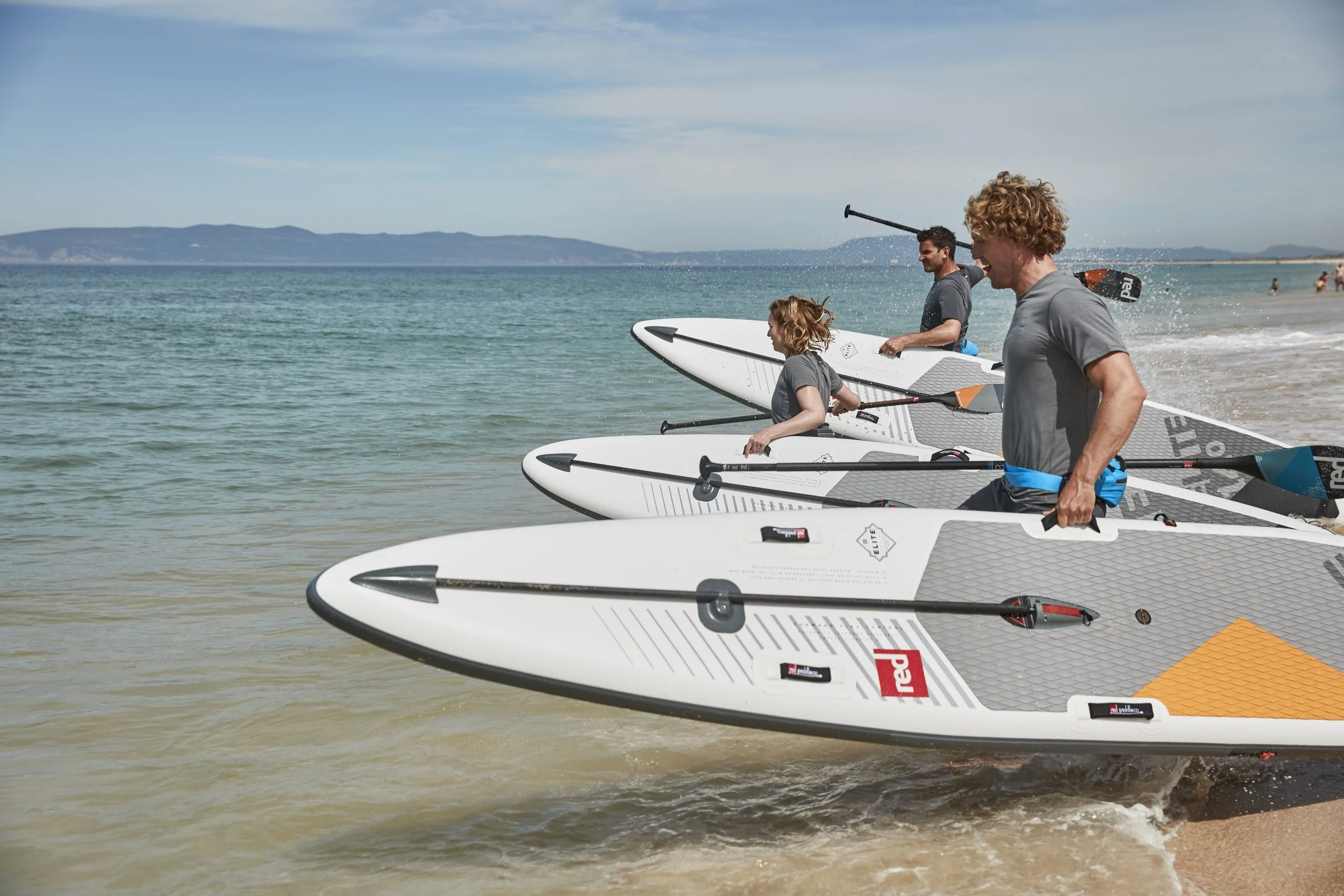 People with paddleboards and paddles at the beach, preparing to go out onto the water.