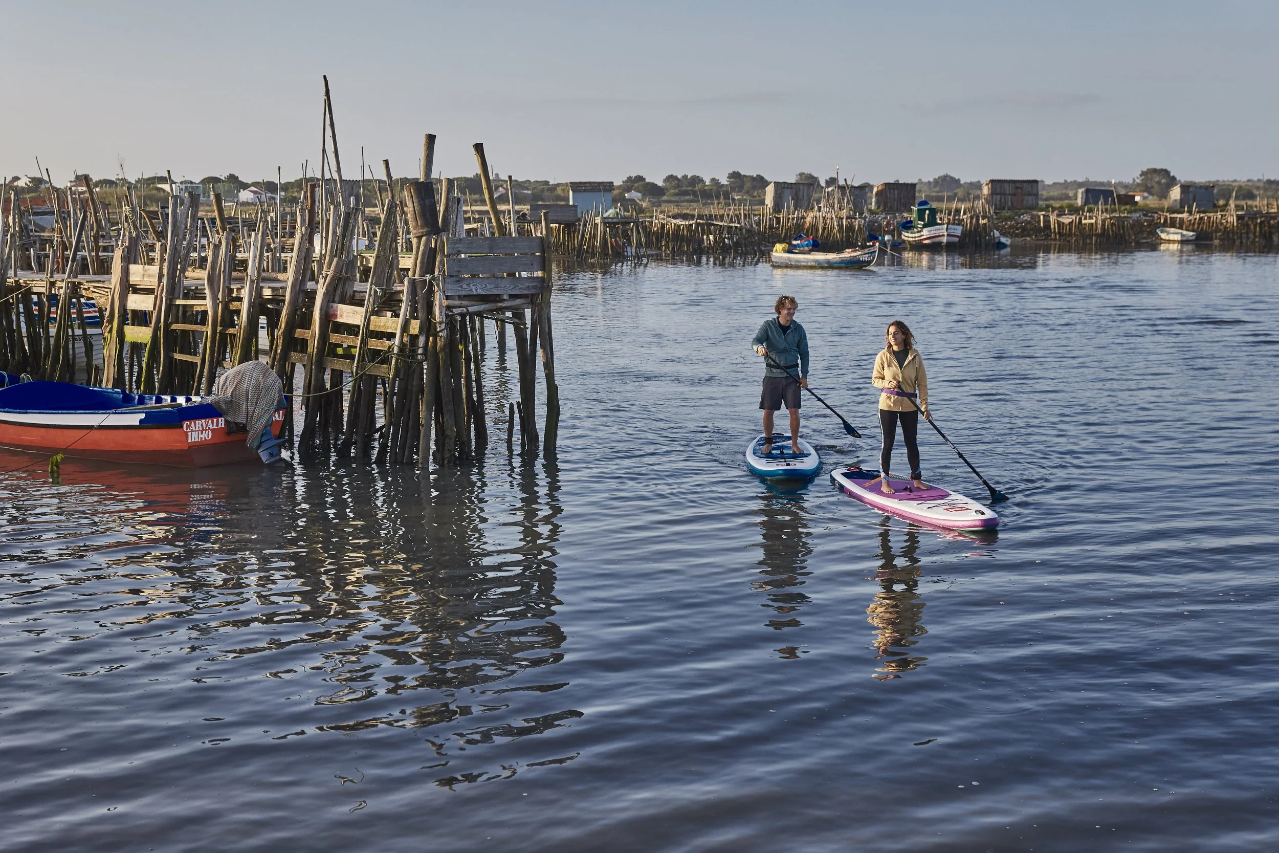 Two people paddleboarding on calm water near a rustic wooden pier with boats and small structures in the background.