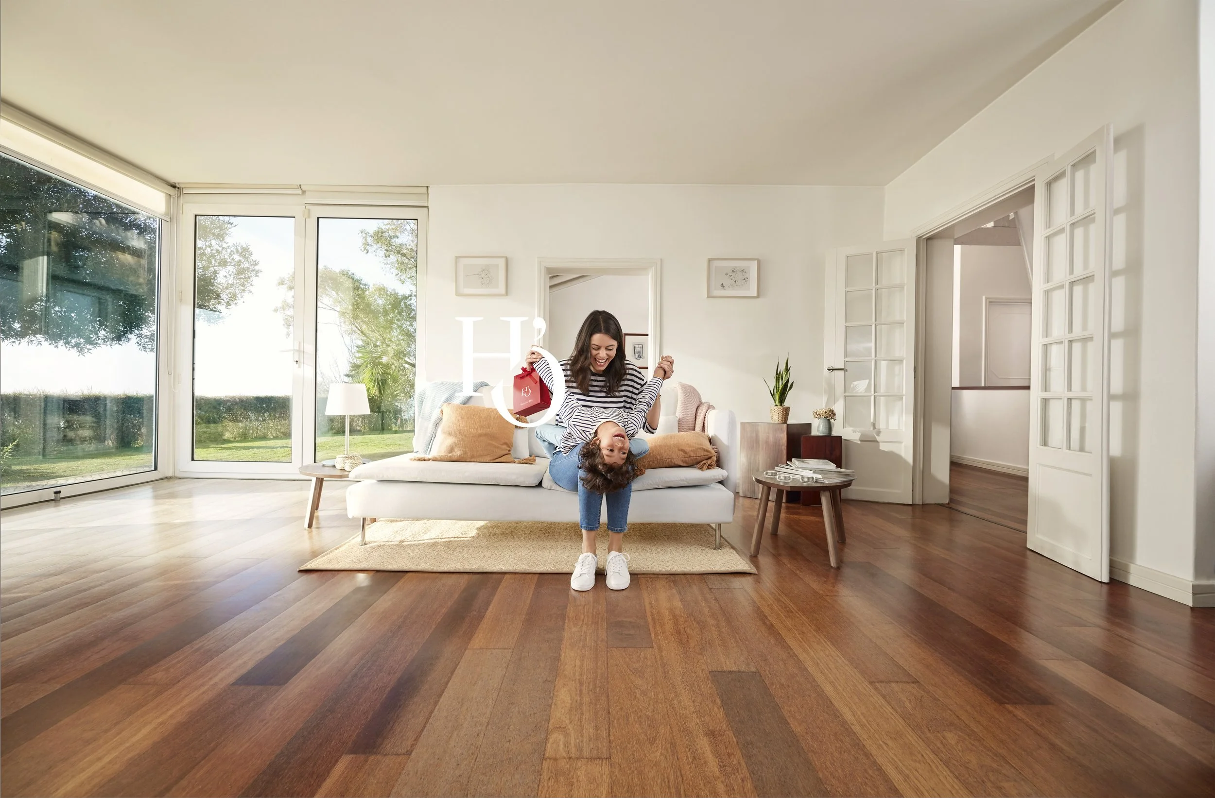 Two women are playfully flipping upside down in a bright, modern living room with wooden flooring and large windows showing a garden outside.