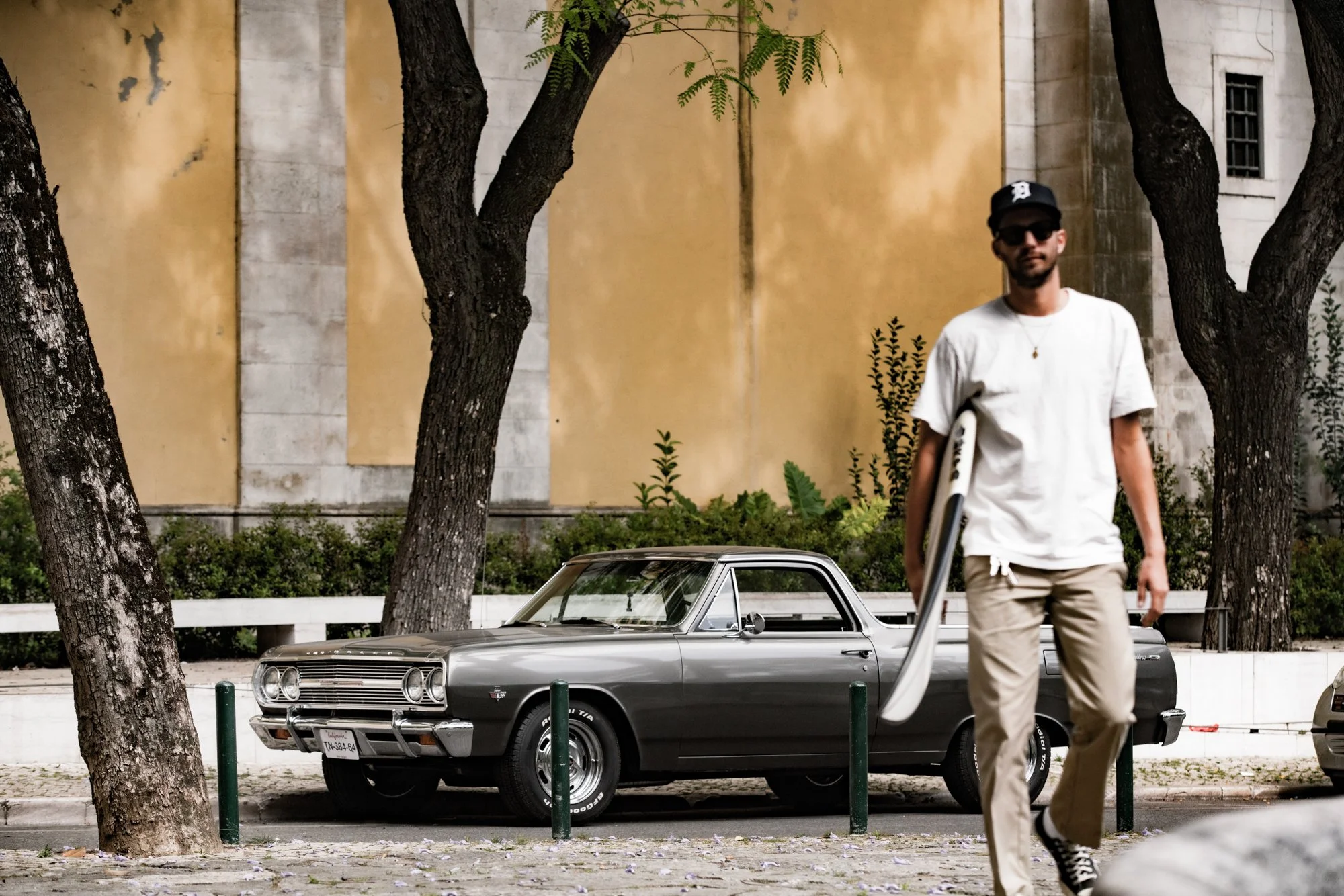 A man wearing sunglasses, a black baseball cap, a white t-shirt, and beige pants walking past a vintage black and silver car parked on the street, with trees and a yellow building in the background.