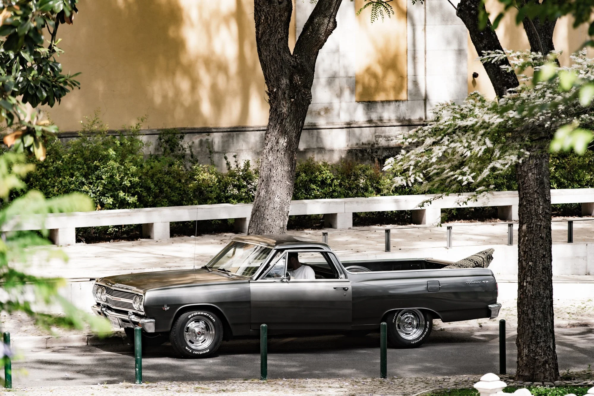 A vintage silver and black car parked on the street in front of a beige wall with trees and white flowering bushes in the background.