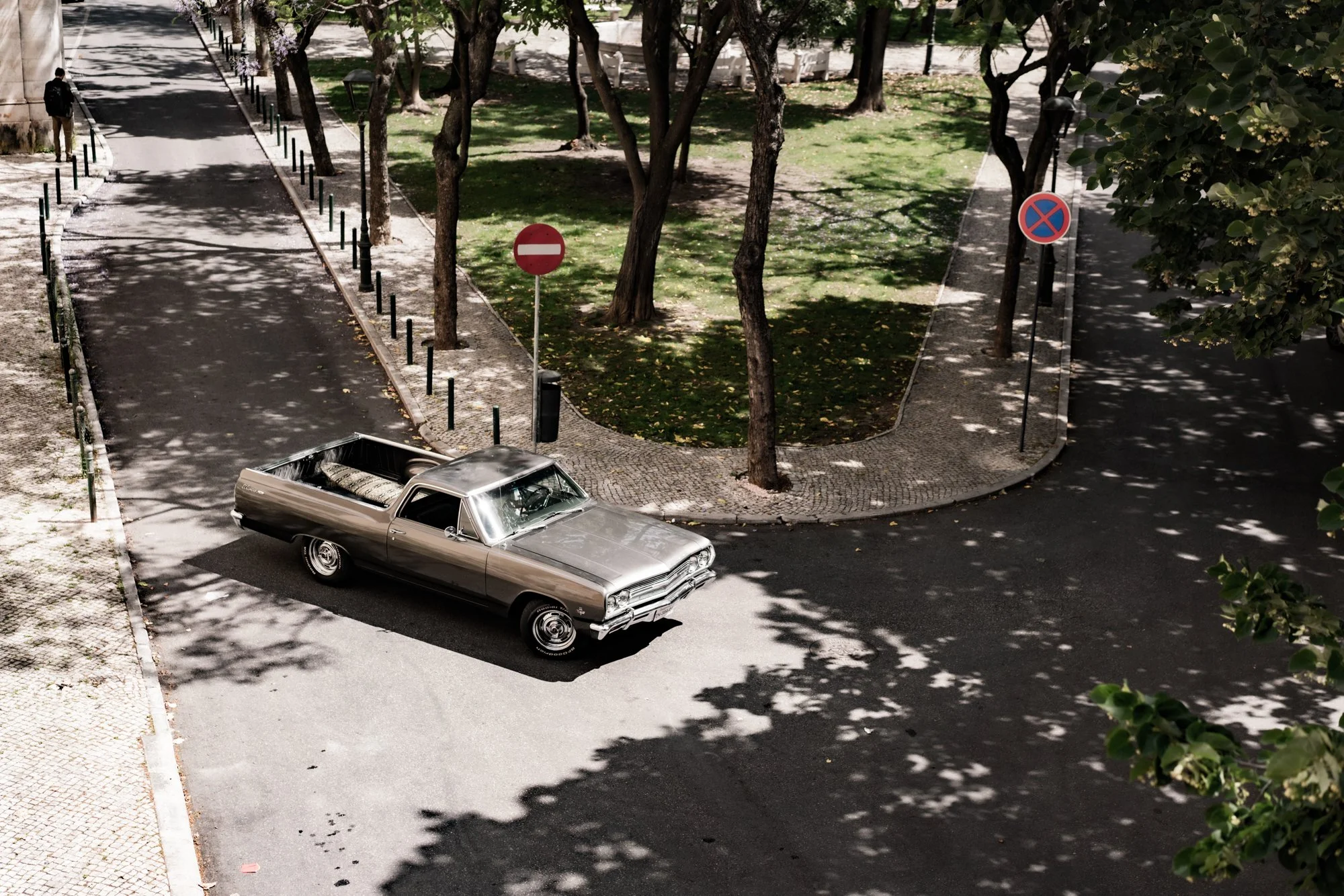 A vintage silver car parked on a street corner near a small park with trees and green grass; there are traffic signs indicating no entry and no parking, and shadows of tree leaves are cast on the street and sidewalk.