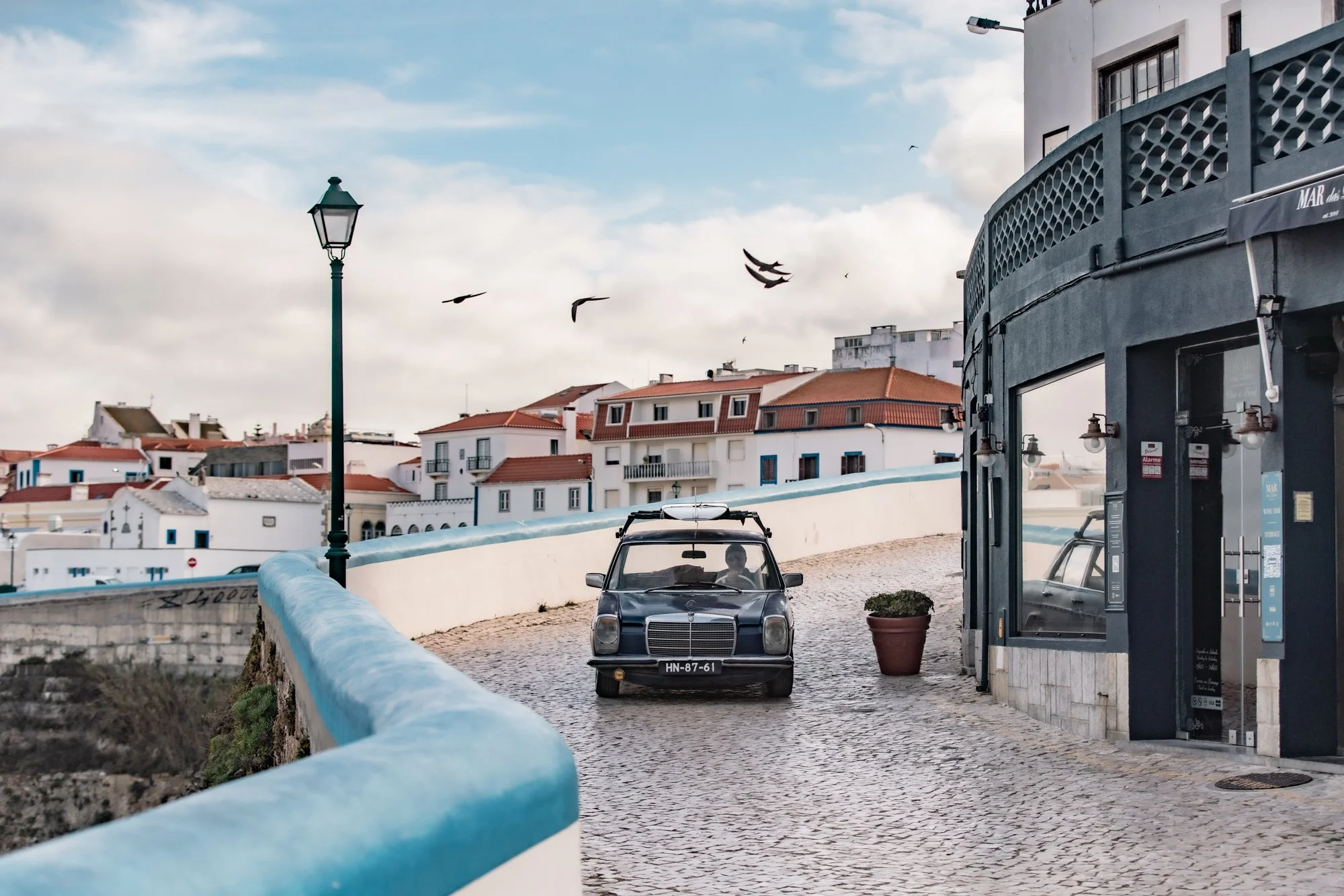 A small cobblestone street on a hill with a vintage black Mercedes-Benz car parked at the top. A green lamppost is beside the street. Residential buildings with white walls and red-tiled roofs are in the background under a cloudy sky, with birds flying.