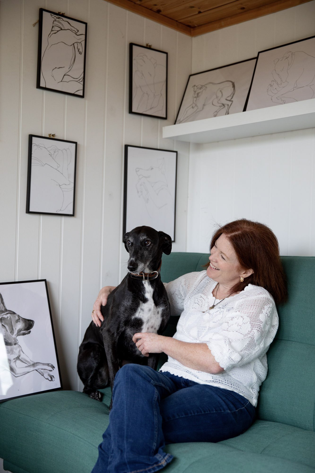 Claudia Dharamshi, female artist with red hair smiling and sitting on a green couch, holding a black and white lurcher, in a room with white paneled walls adorned with framed line art drawings of dogs.