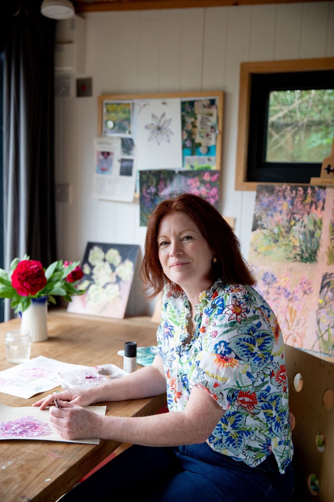 Claudia Dharamshi, female artist with with red hair and a floral shirt sitting at a wooden table, working on watercolor paintings with pink flowers, in an art studio with paintings on the wall and a window showing greenery outside.