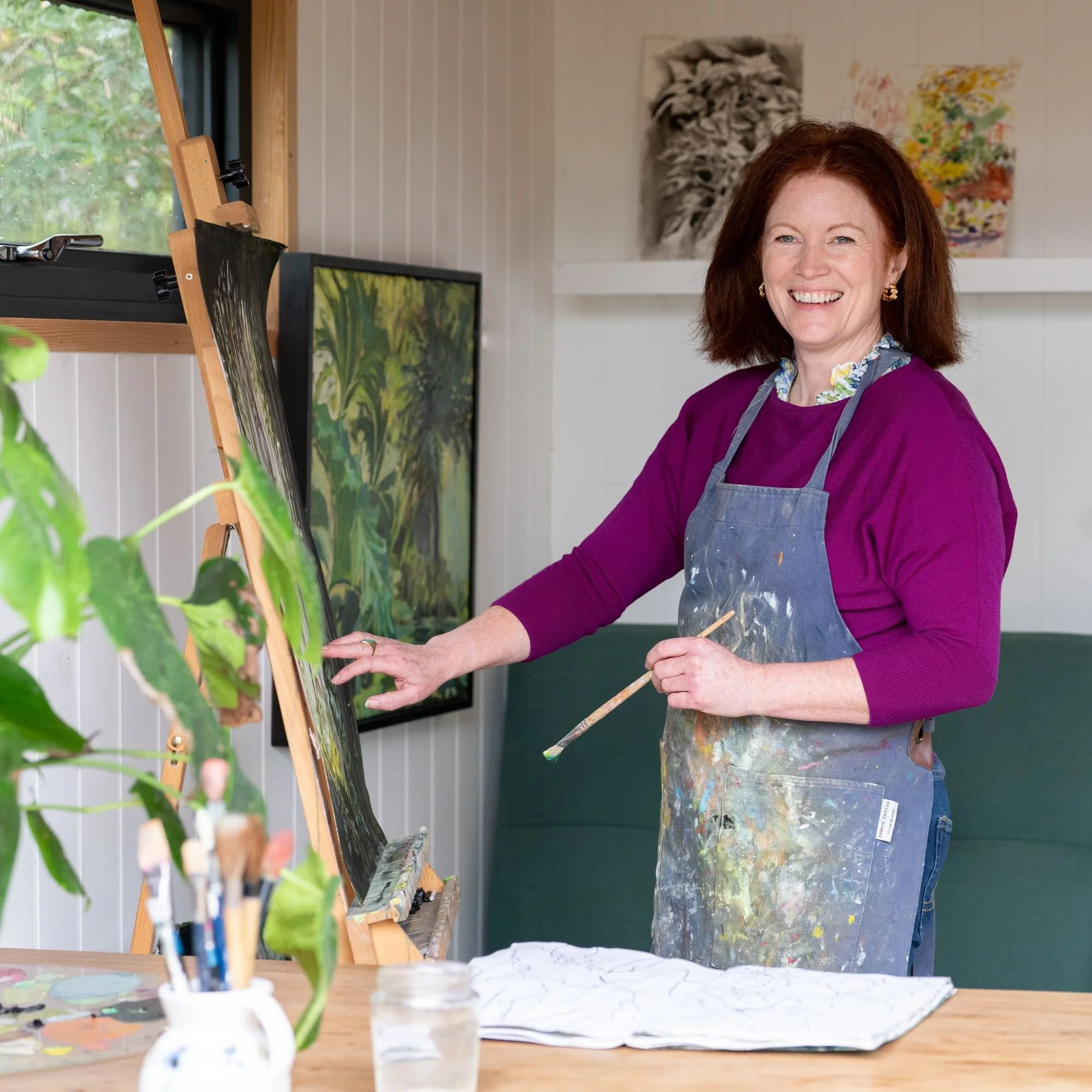 Claudia Dharamshi, a woman with red hair and a purple sweater, wearing a paint-splattered apron, smiling while painting a jungle scene at an easel in her art studio.