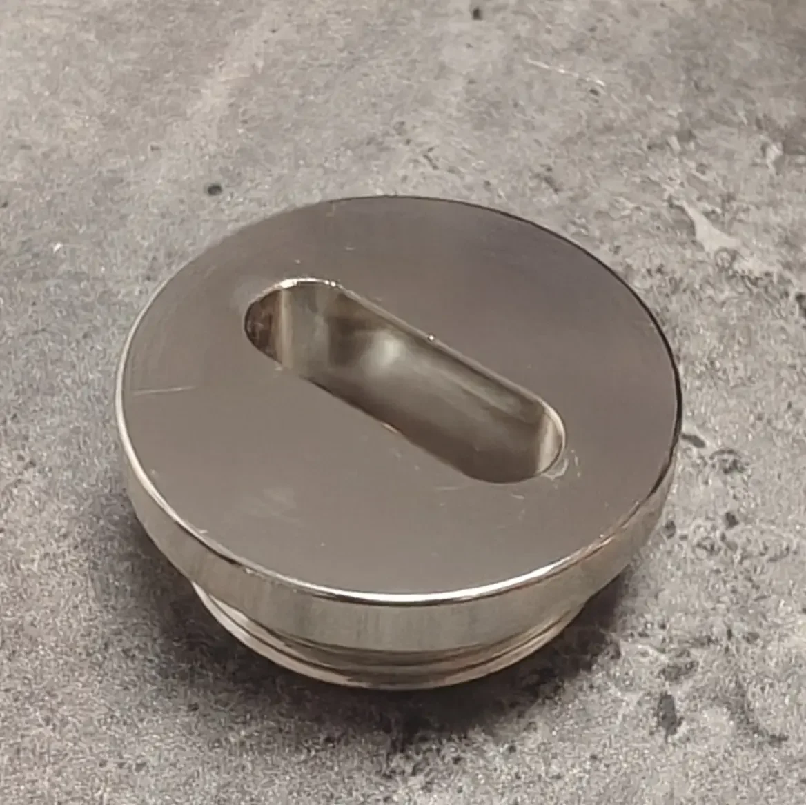 A shiny, metallic, oval-shaped speaker with a horizontal slot on top, resting on a rough, concrete surface.
