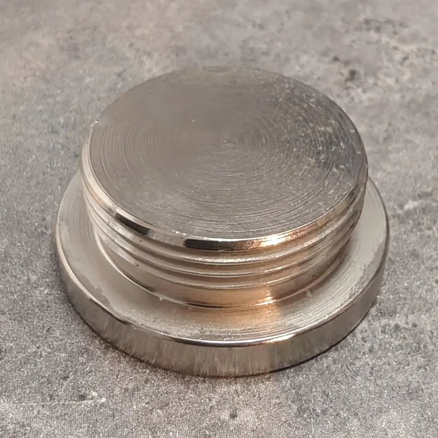 Close-up of a metal screw top, showing the circular grooves on the top surface and a threaded ring around the side, resting on a textured gray surface.