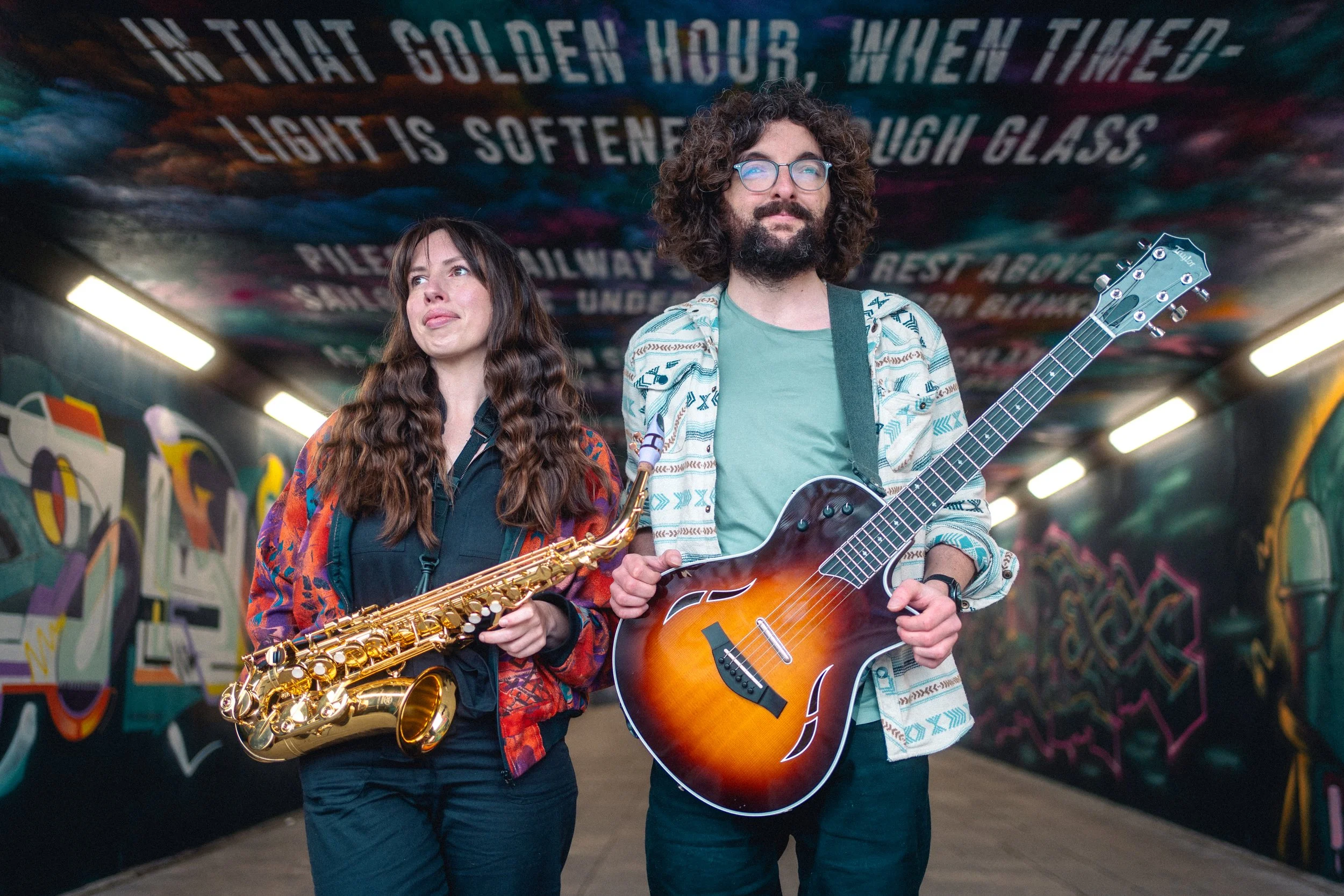 A man and woman standing in an urban tunnel with colorful graffiti on the walls and a painted message on the ceiling. The woman holds a saxophone, and the man holds an acoustic guitar. The ceiling message reads: 'In that golden hour, when timed light is softened enough to laugh glass.'