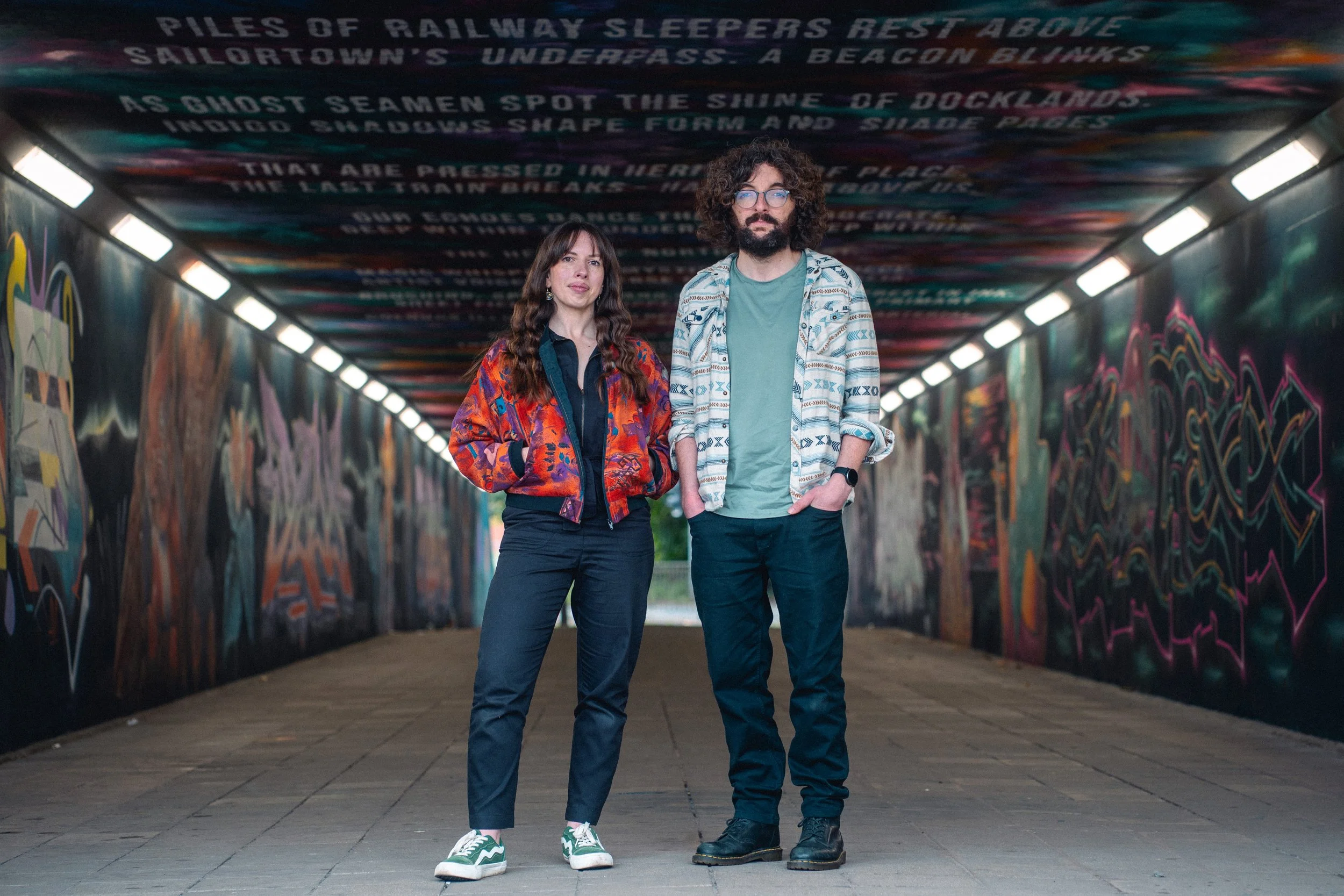 A woman and a man standing in a tunnel with colorful graffiti and words written on the ceiling.