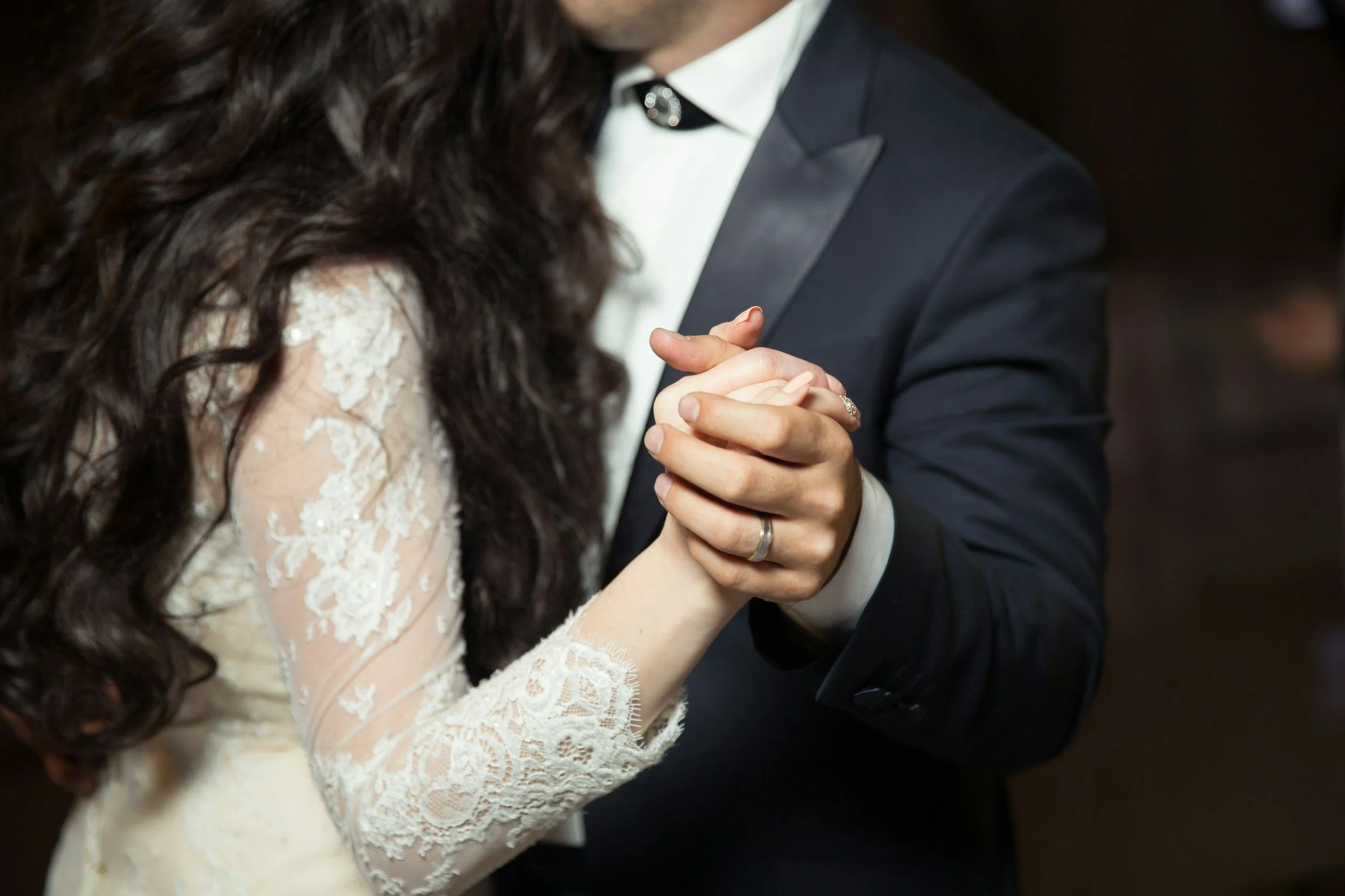 A bride and groom are dancing at their wedding, holding hands with wedding rings visible on their fingers. The bride is wearing a lace wedding gown with long sleeves, and the groom is dressed in a dark suit with a white shirt.