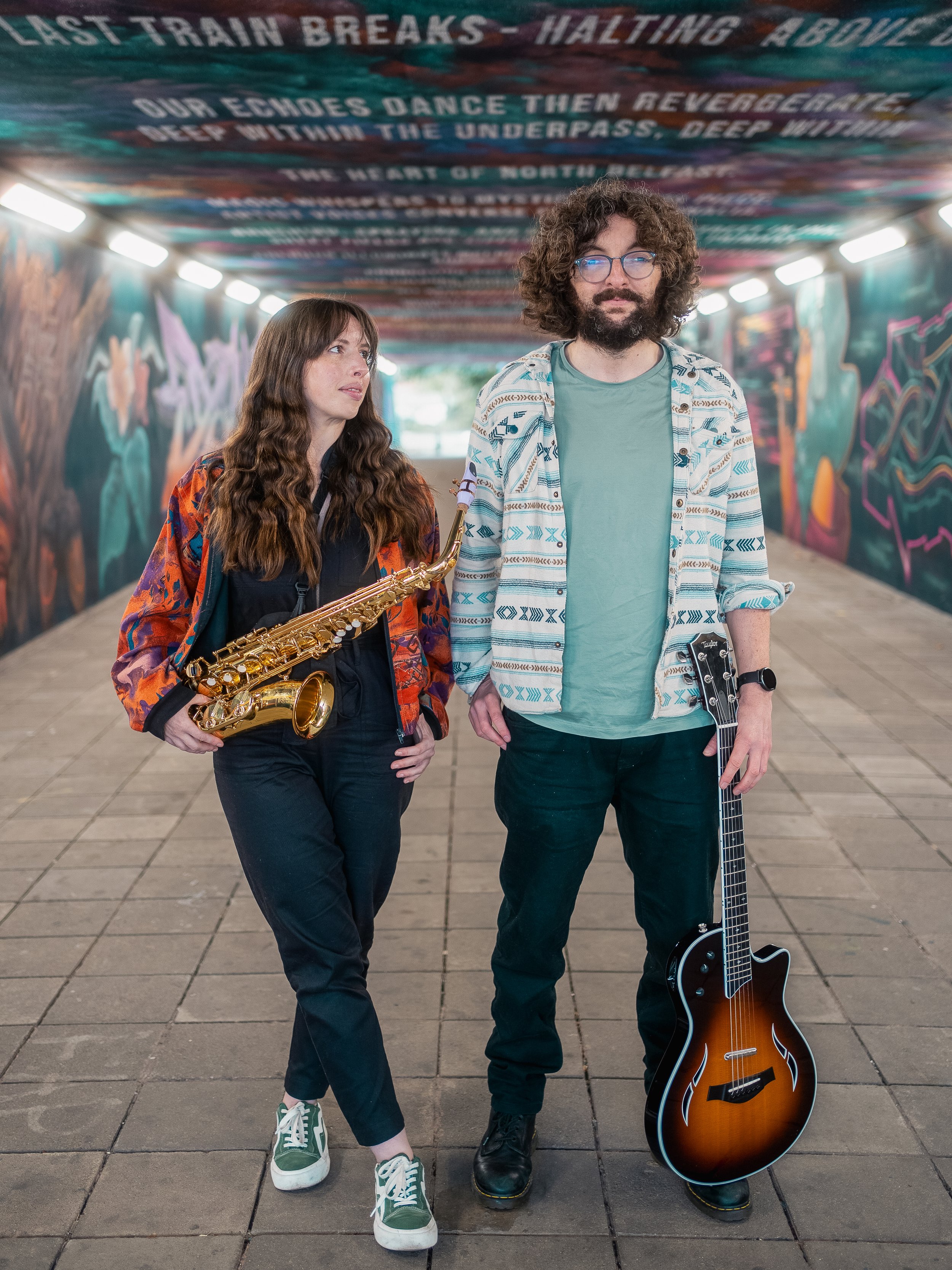 Two musicians walking under a colorful graffiti-covered underpass. The female musician on the left has long wavy brown hair and is holding a saxophone. The male musician on the right has curly hair, glasses, a beard, and is holding an electric guitar. The ceiling of the underpass has painted lyrics about train breaks and dancing.