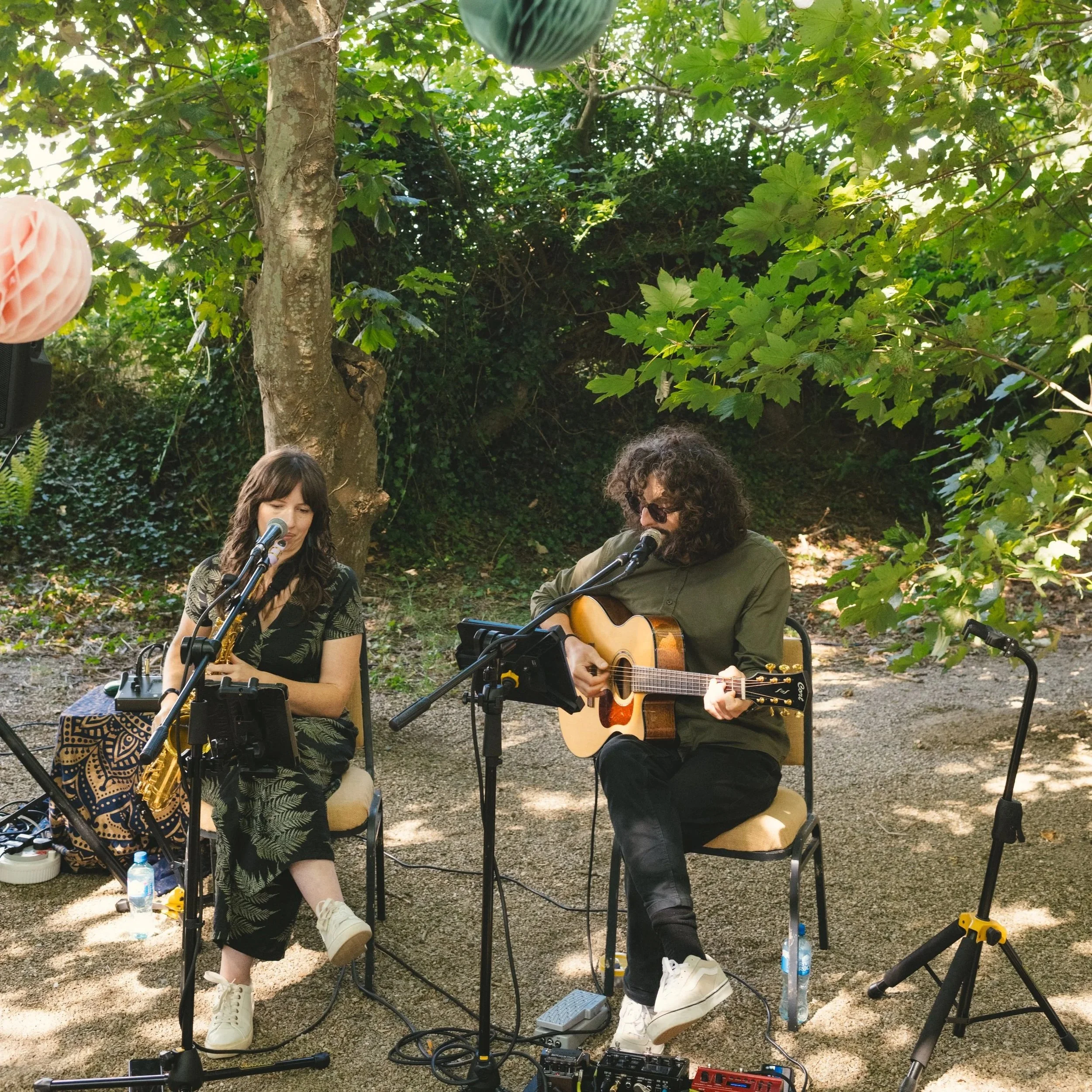 Two musicians performing outdoors in a lush green garden. One woman with long dark hair playing a saxophone, and a man with curly hair wearing glasses playing an acoustic guitar. They are seated in front of microphones with sound equipment around them.