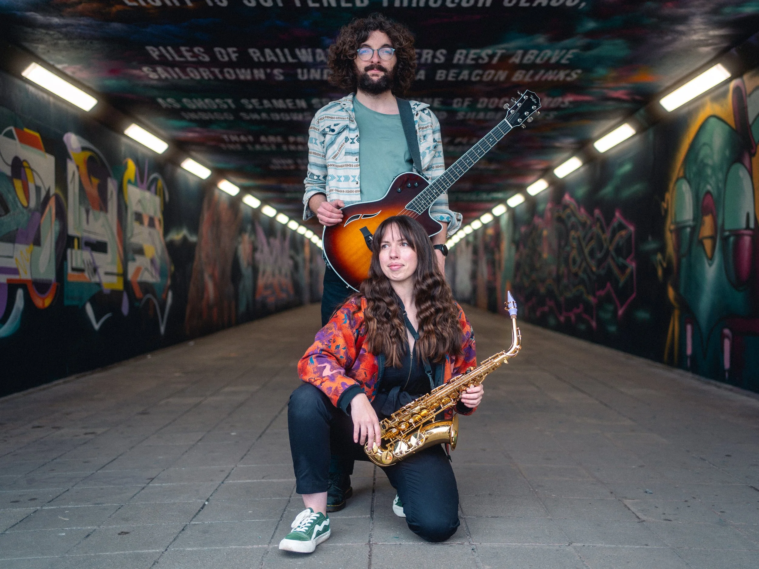 A man with curly hair, glasses, and a beard holding an electric guitar, standing in front of a woman with long hair holding a saxophone, with colorful graffiti art on the walls of an underpass.