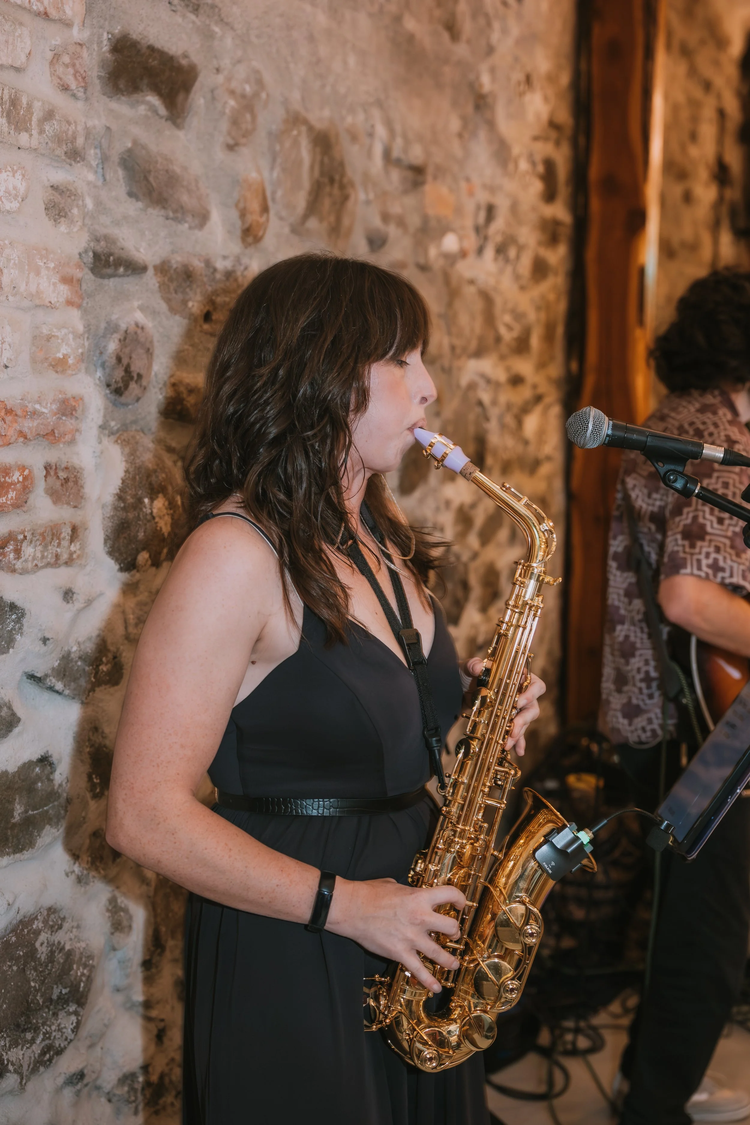 A woman with shoulder-length wavy brown hair playing a gold saxophone, wearing a black sleeveless top and a black bracelet, standing against a rustic brick wall, with a microphone nearby.