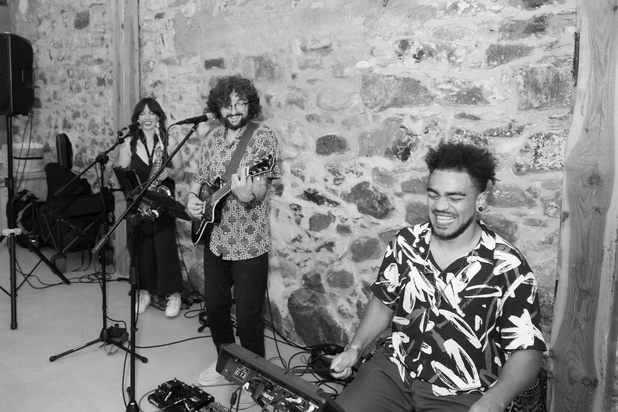A group of musicians performing in a rustic venue with a stone wall background. One man with curly hair and glasses is playing an electric guitar and singing into a microphone. A woman with dark hair in braids is smiling in the background, standing near a keyboard. Another man with spiky hair, wearing a patterned shirt, is playing a keyboard and smiling. Various audio equipment and cables are visible.