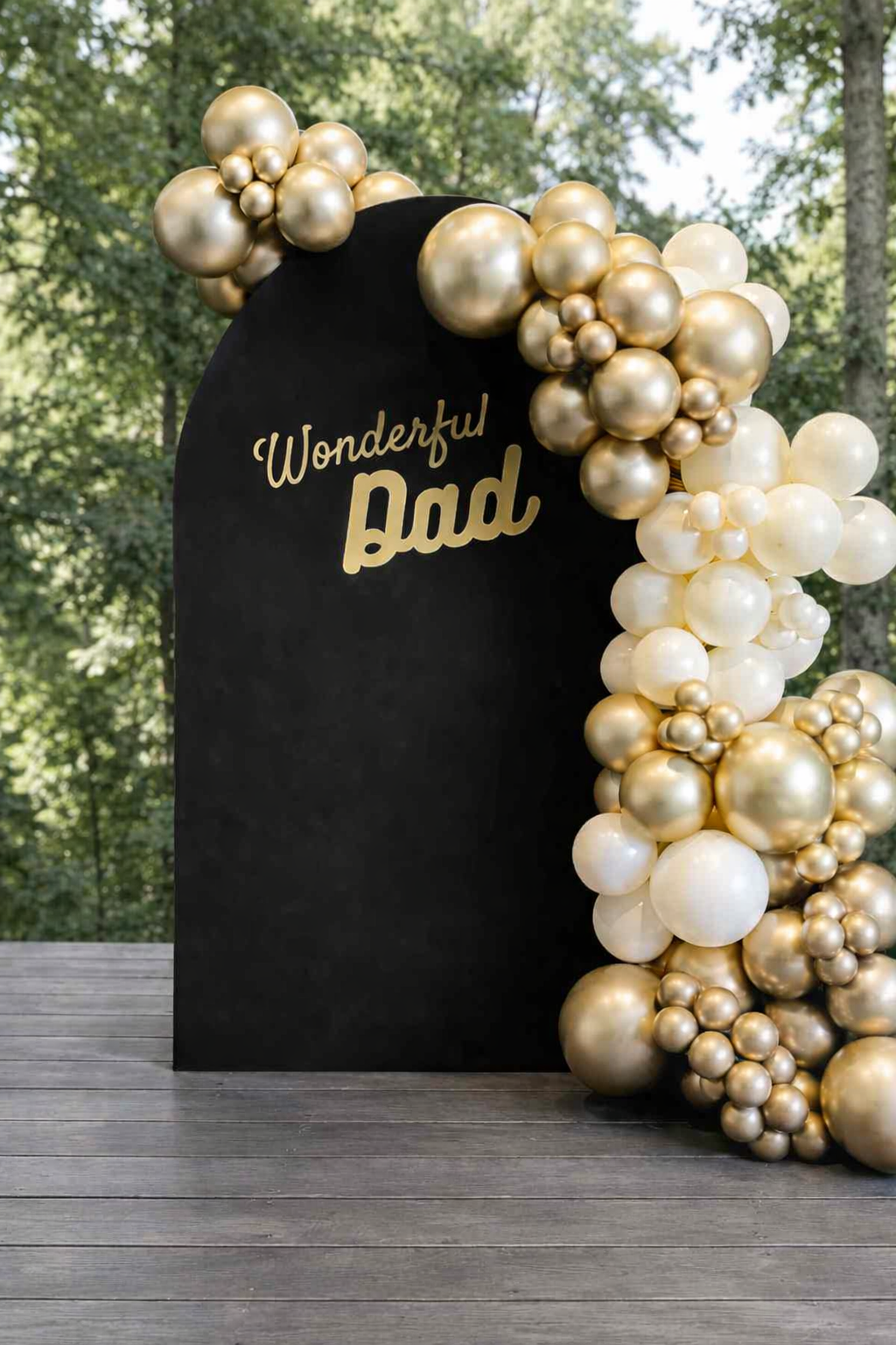 A black standing board with the words 'Wonderful Dad' written in gold, surrounded by gold, white, and pearl-colored balloons arranged in a decorative cluster on an outdoor wooden deck with trees in the background.