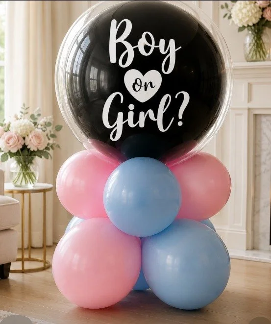 A cluster of pink and blue balloons with a large black and clear balloon on top that reads "Boy or Girl?" in white script. The background shows a living room with a fireplace and a side table with flowers.